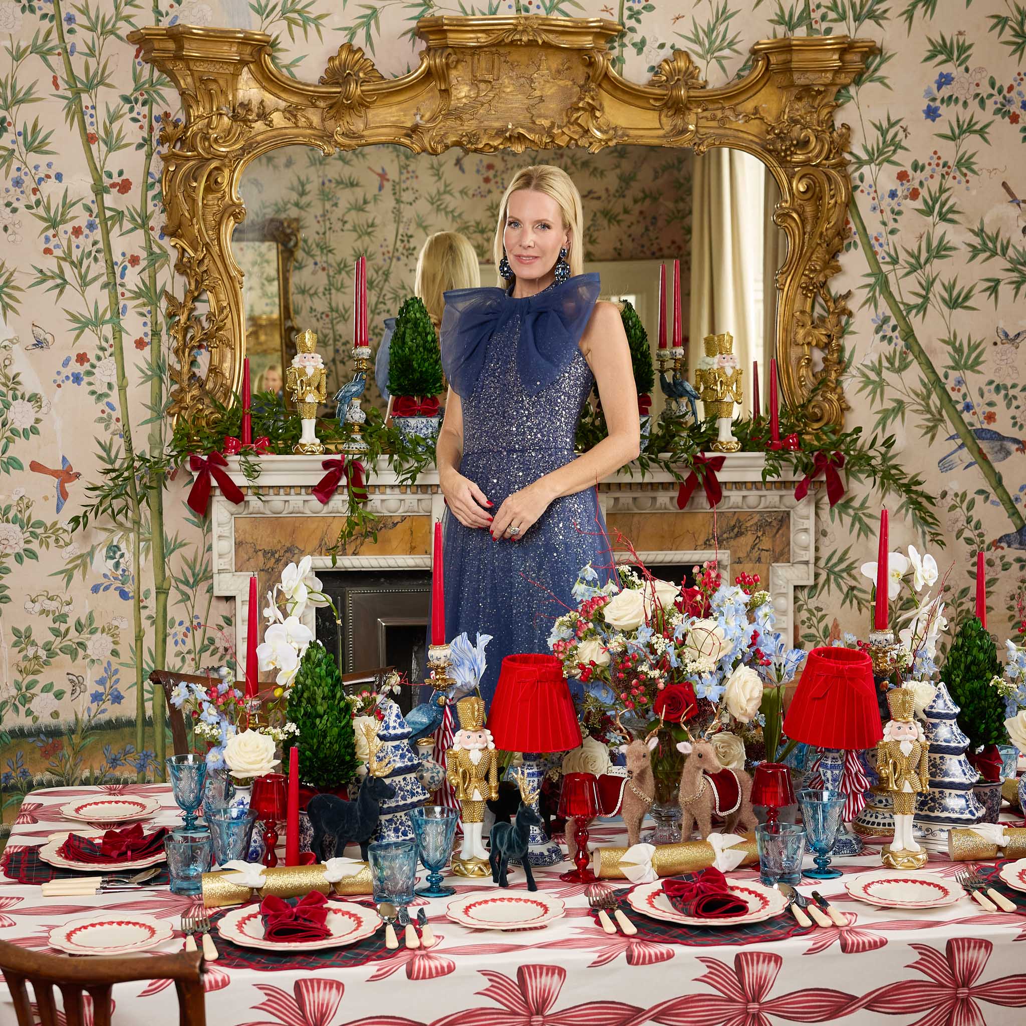 Alice in a blue dress standing in a christmas decorated room with a large mirror and festive table setting.