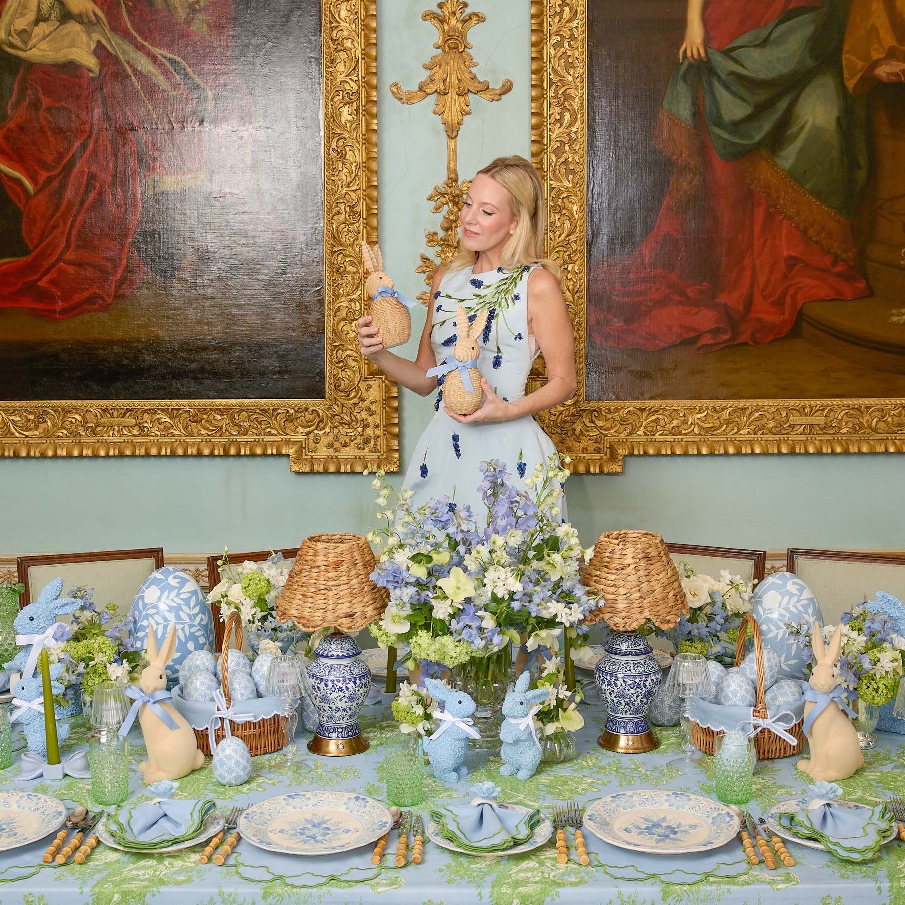 Woman standing in a room with classical paintings, holding a bunny , with a decorated table in front of her.