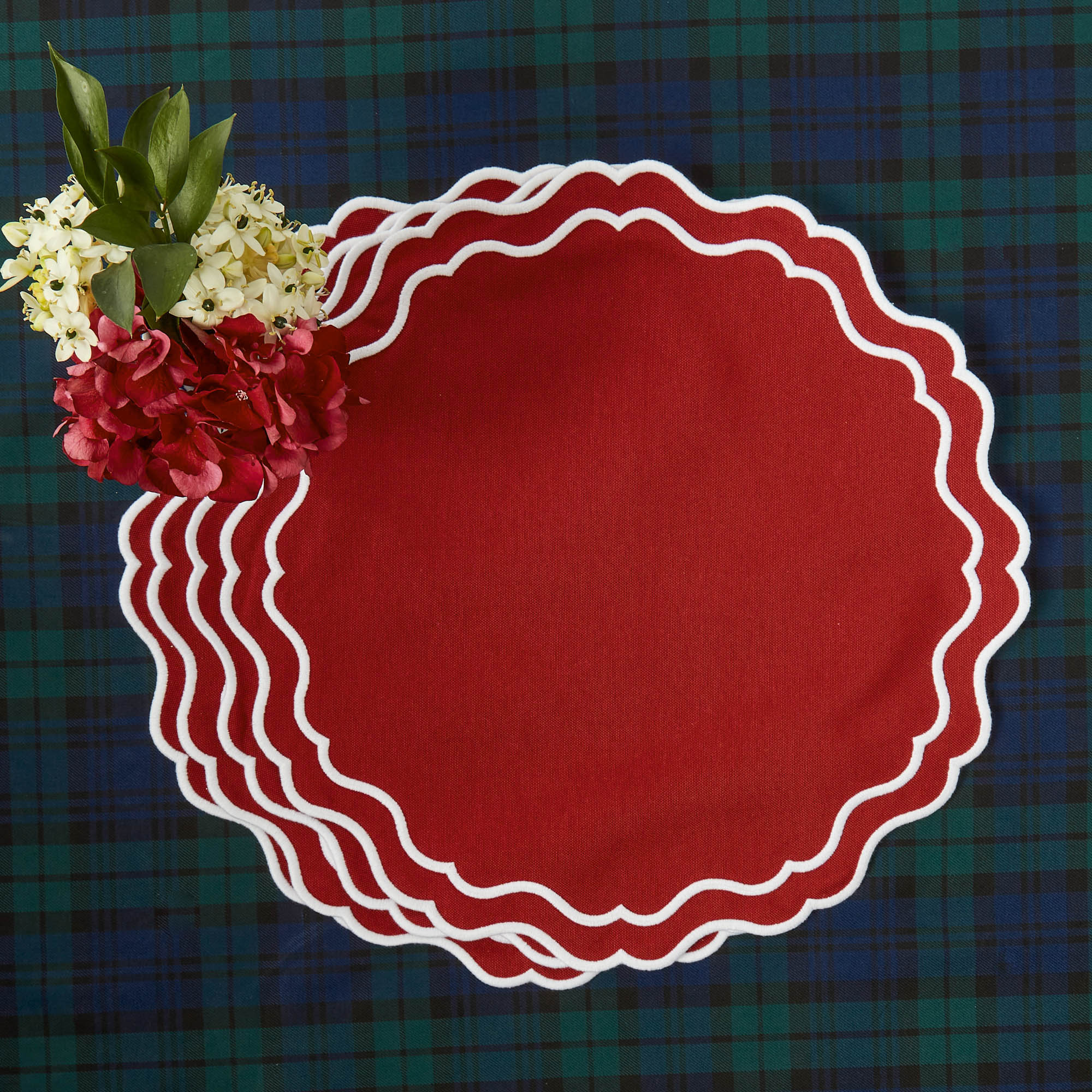 A set of four round, red placemats with a double embroidered edge on a scolloped style, displayed on a background of a tartan tablecloth with a small bouquet of flowers on top.