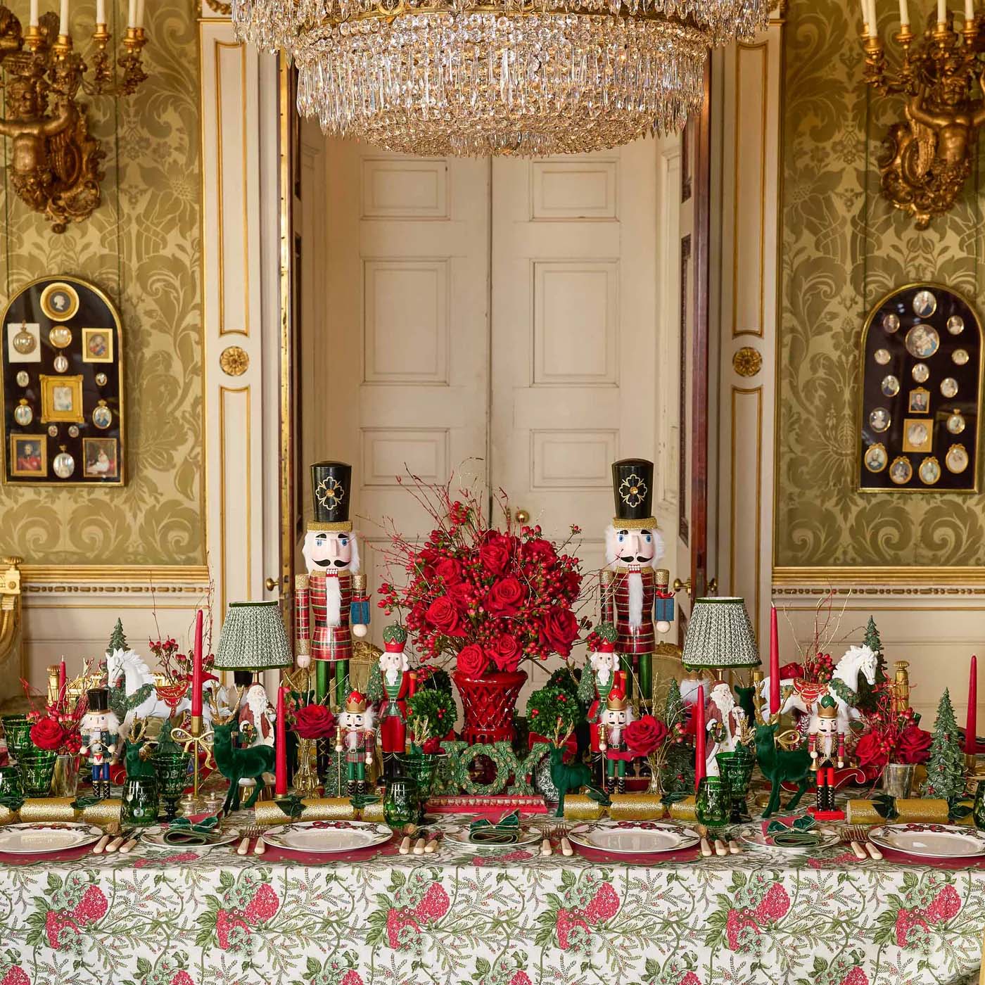 Decorative table setting with red flowers and greenery in a grand room with chandeliers.
