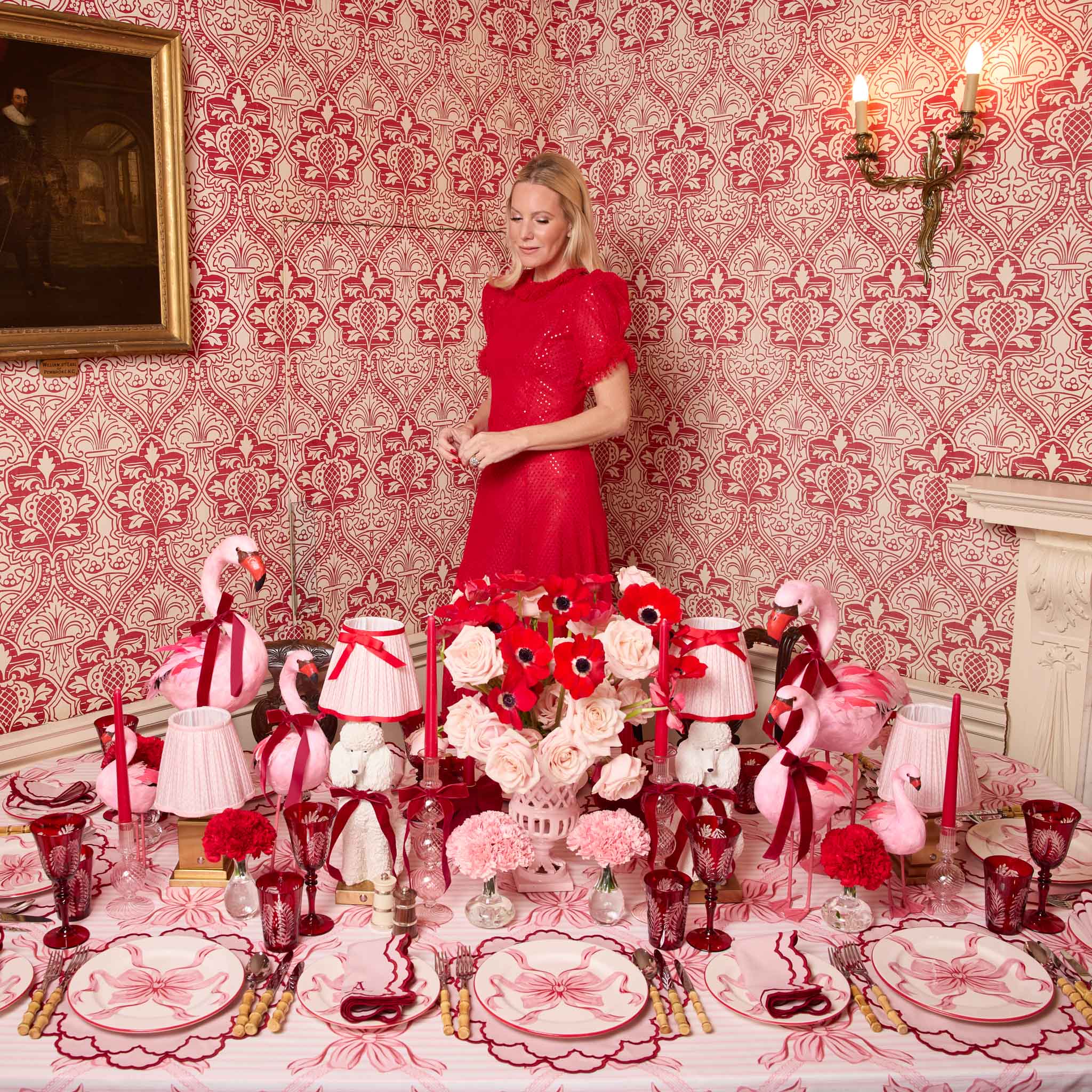 Woman in a red dress standing behind a elaborately decorated table with floral arrangements and flamingo decorations against a patterned wall. for valentines day.