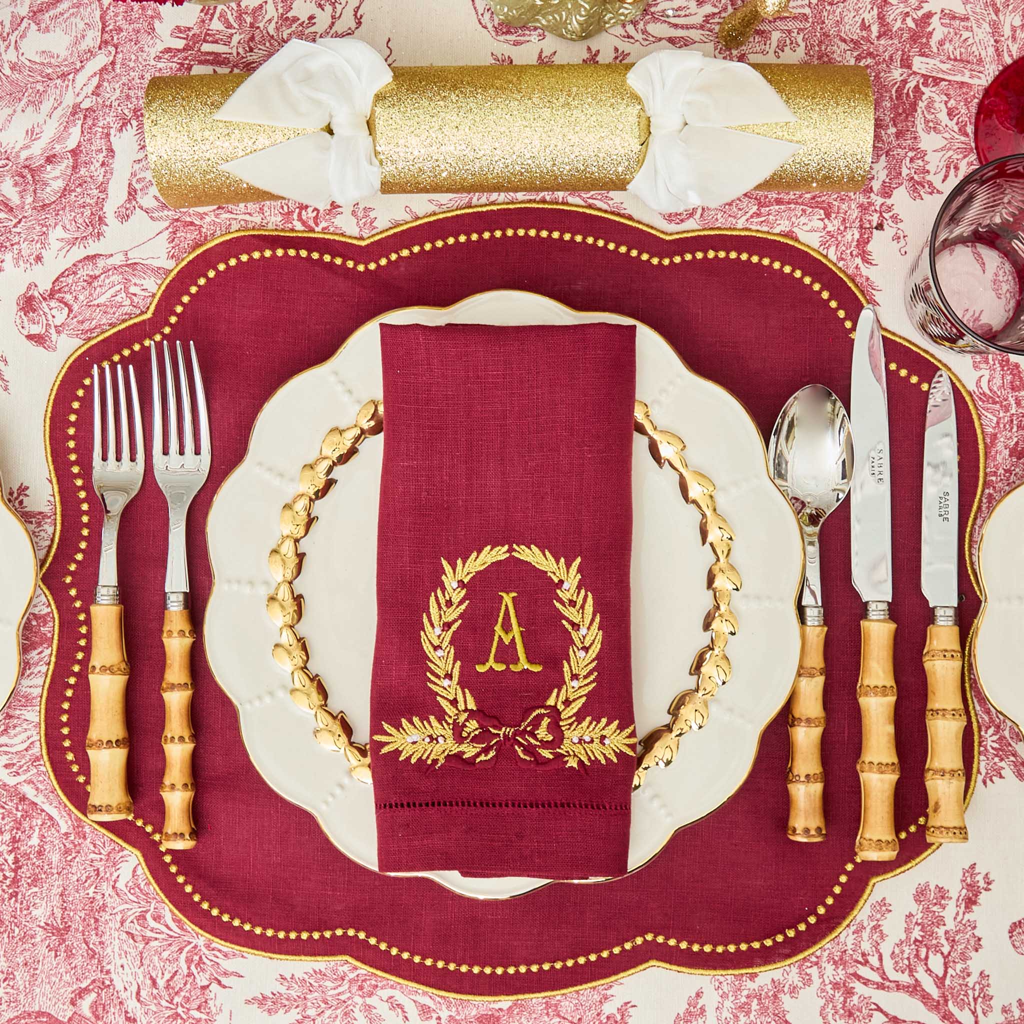 Decorative table setting with gold cutlery, red napkin with monogram, and white plates on a patterned tablecloth.