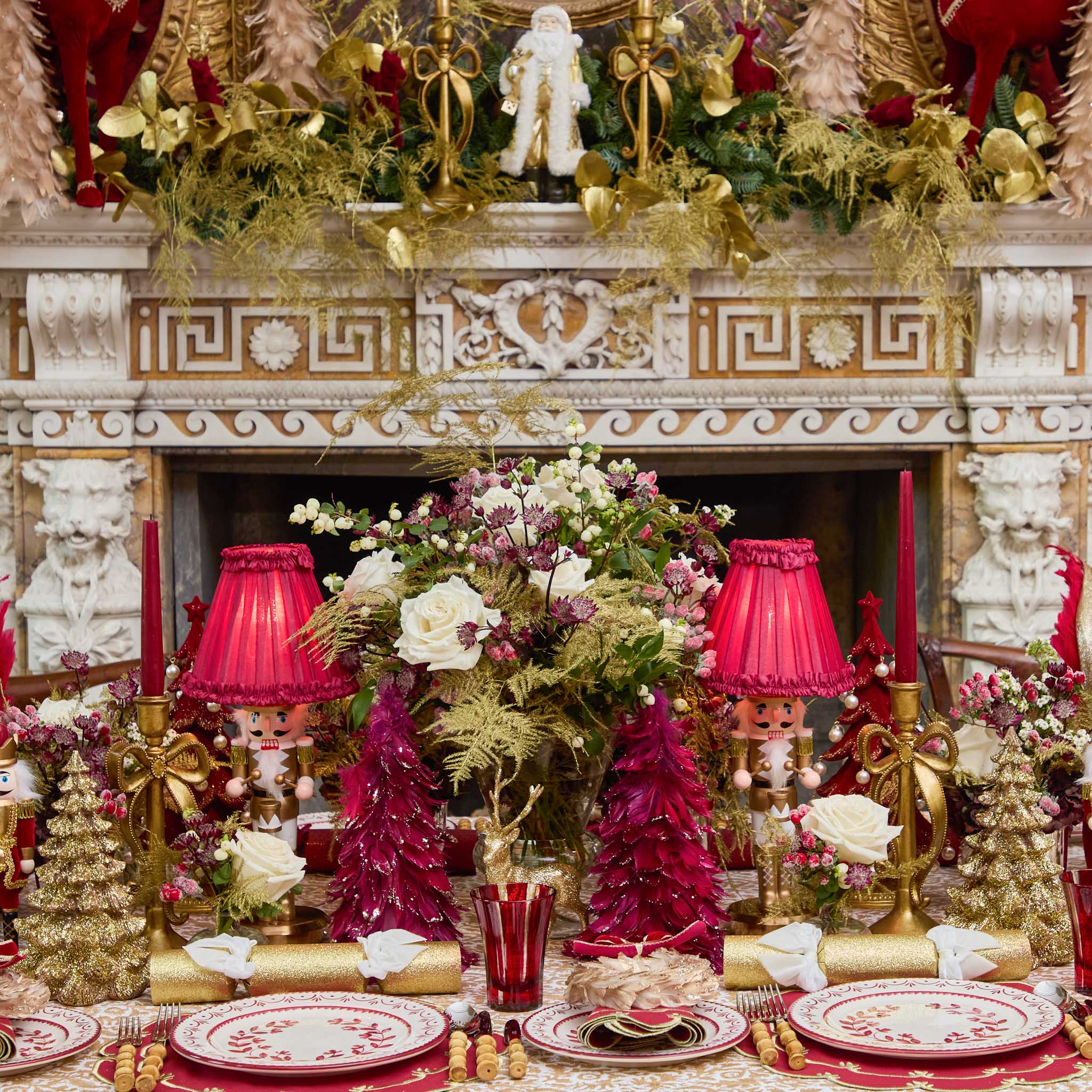 Decorative Christmas table setting with floral arrangements, red lamps, and festive decorations in front of a decorated fireplace.