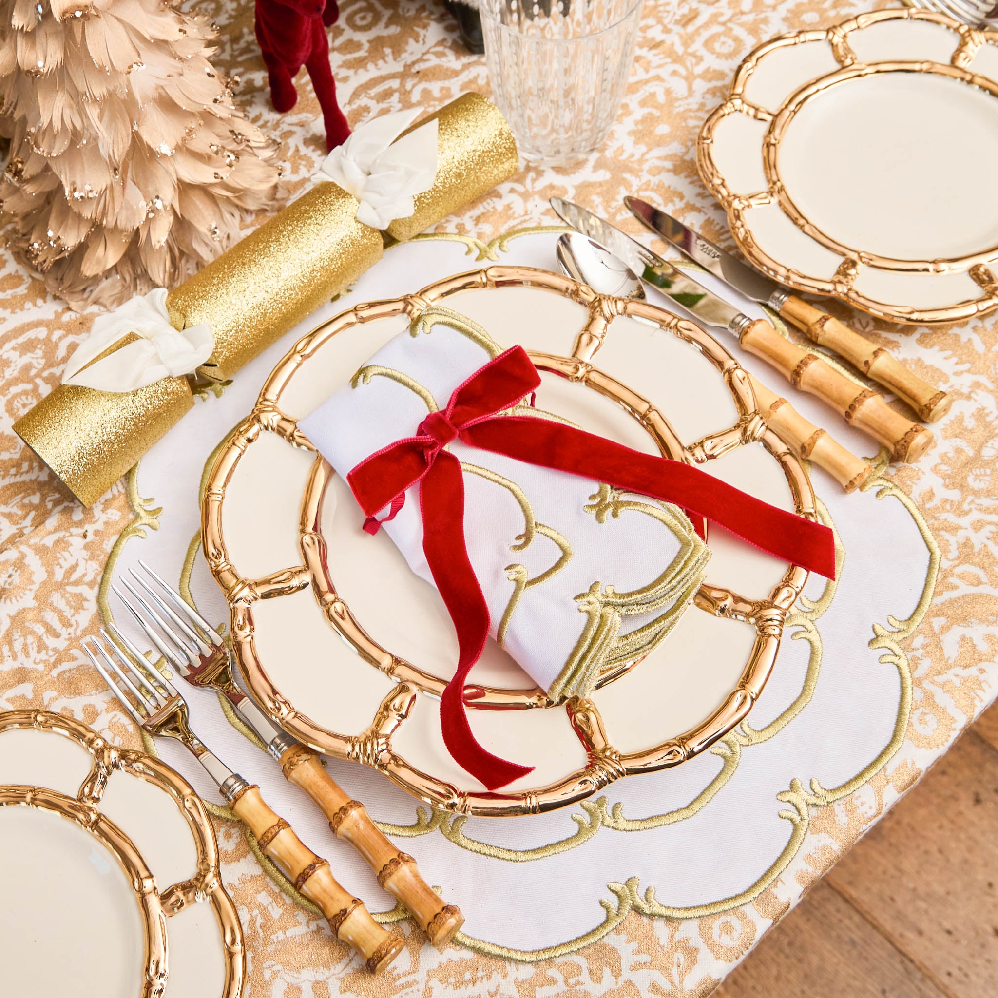 Decorative table setting with gold-rimmed plates, red ribbons, and cutlery on a patterned tablecloth.