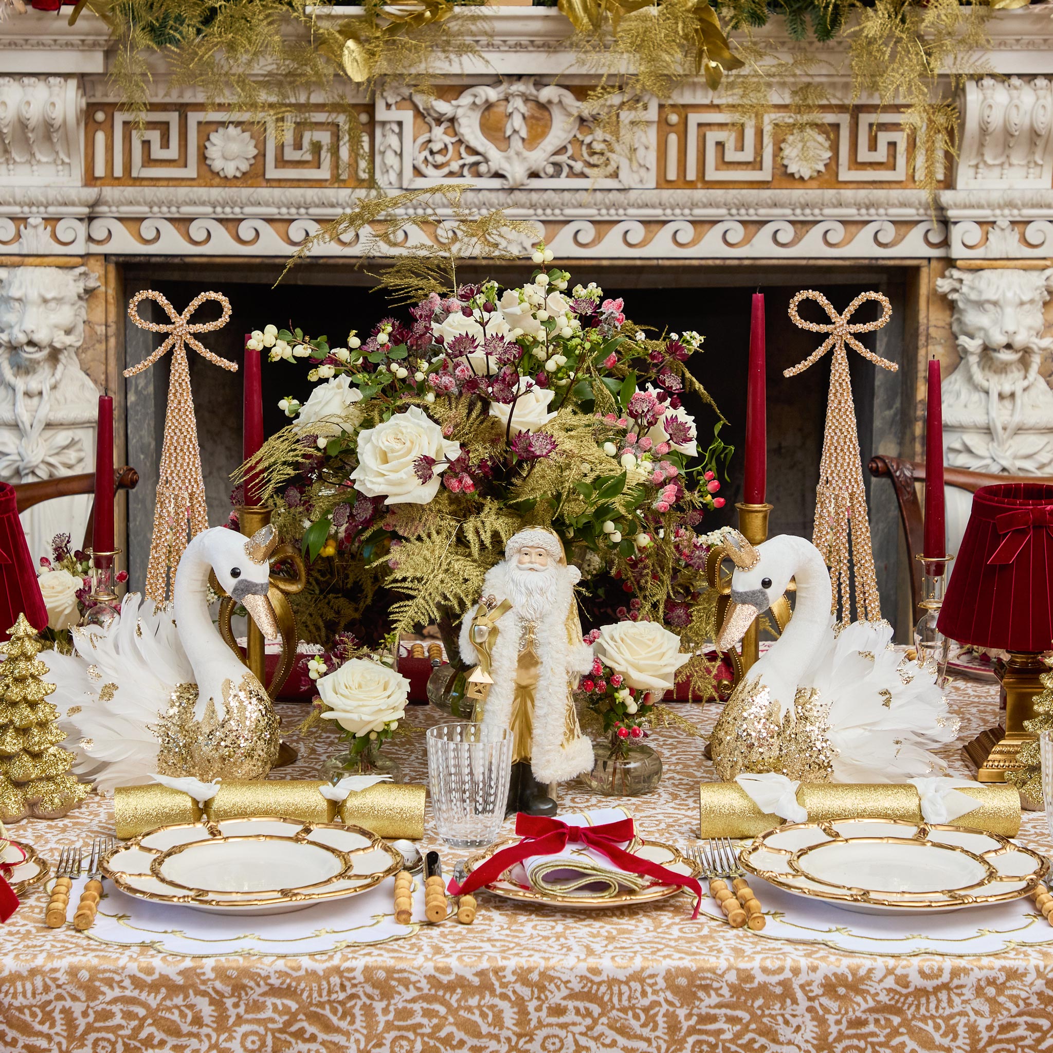 Decorative Christmas table setting with swans, Santa figure, and floral arrangement in front of a fireplace.