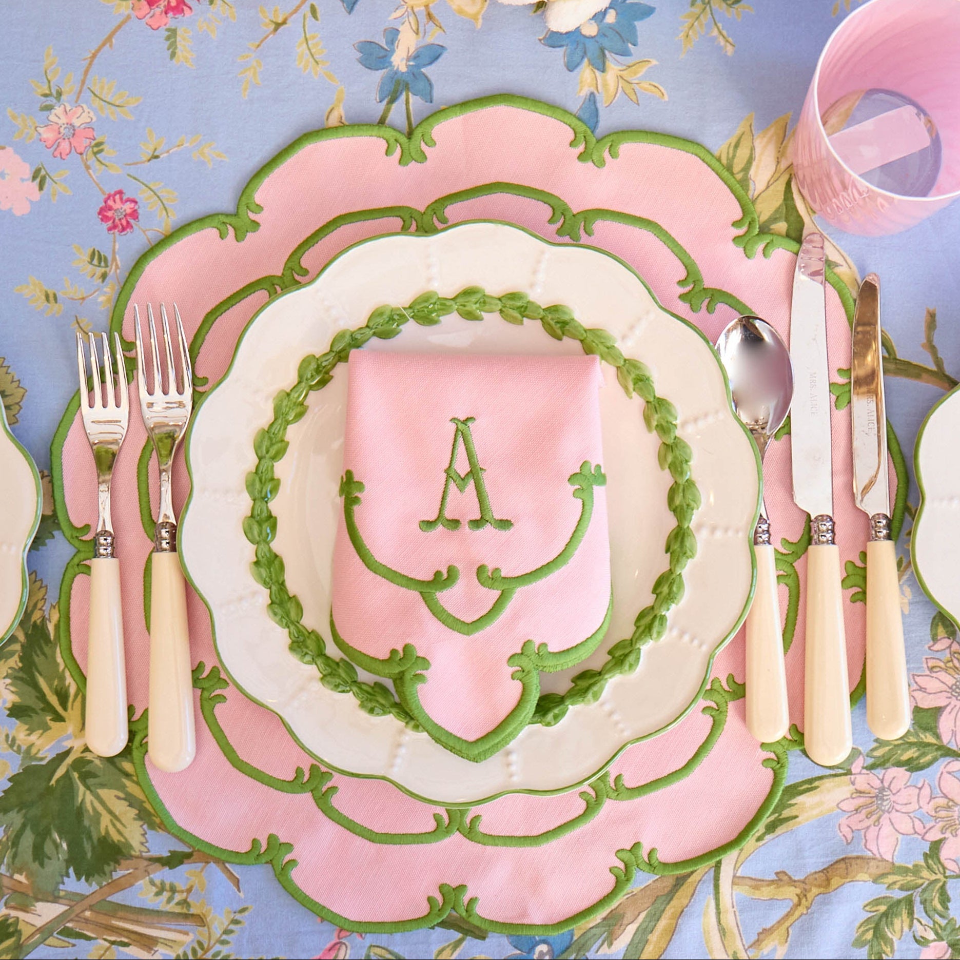 Decorative table setting with pink and green plates, cutlery, and floral tablecloth.