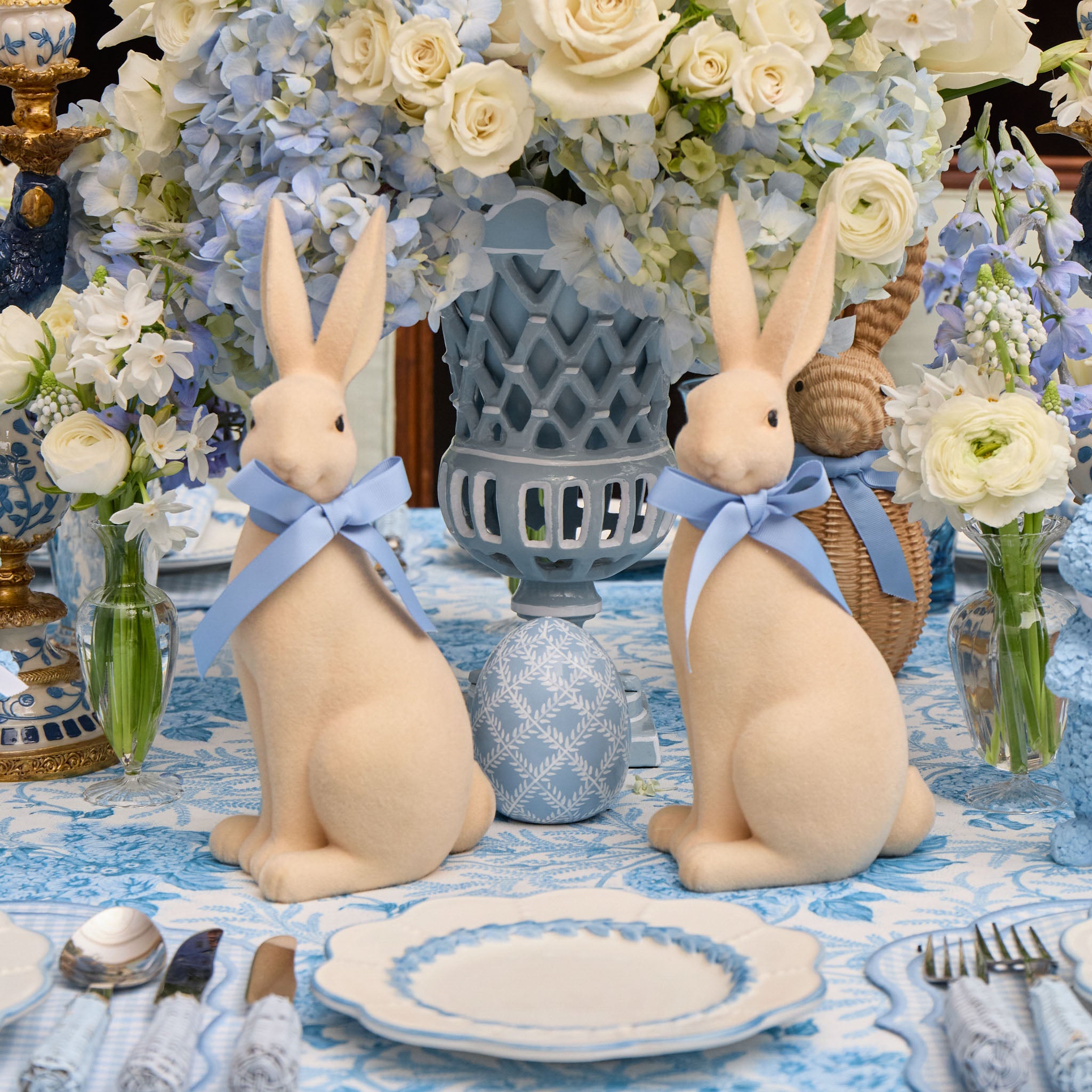 Two decorative rabbits with blue ribbons on a table setting with flowers and tableware.
