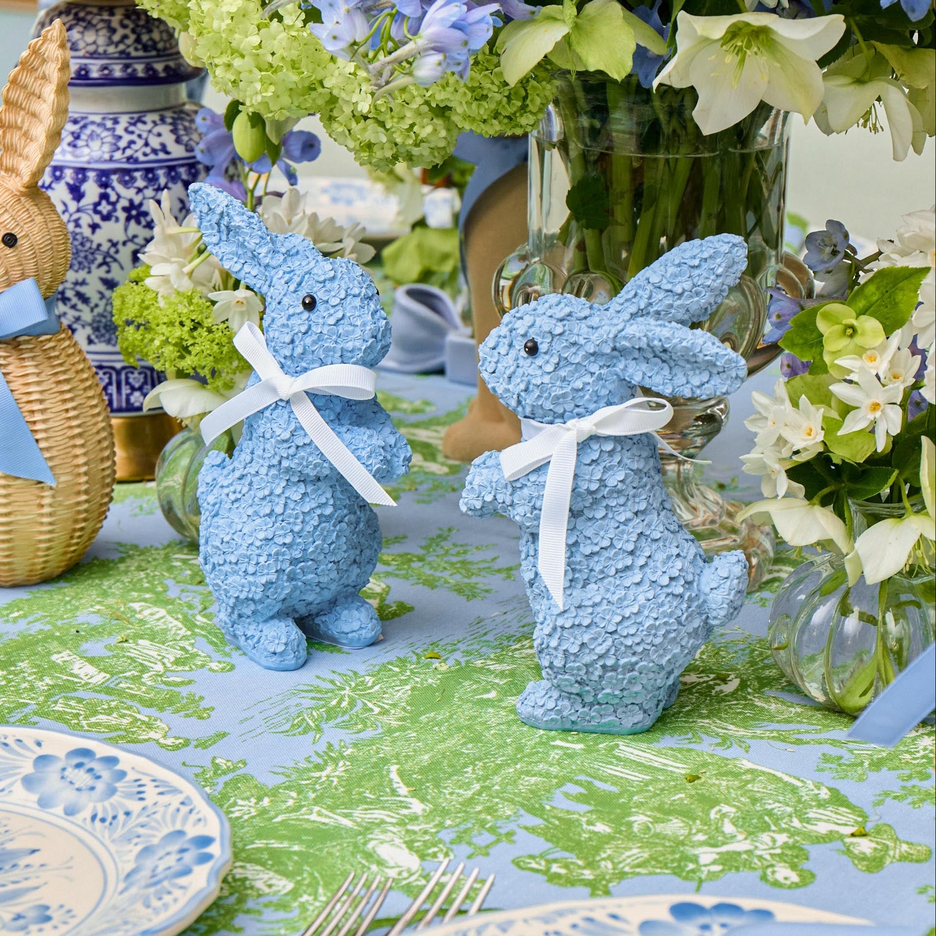 Decorative blue bunny figurines on a table with floral arrangements and patterned tablecloth.