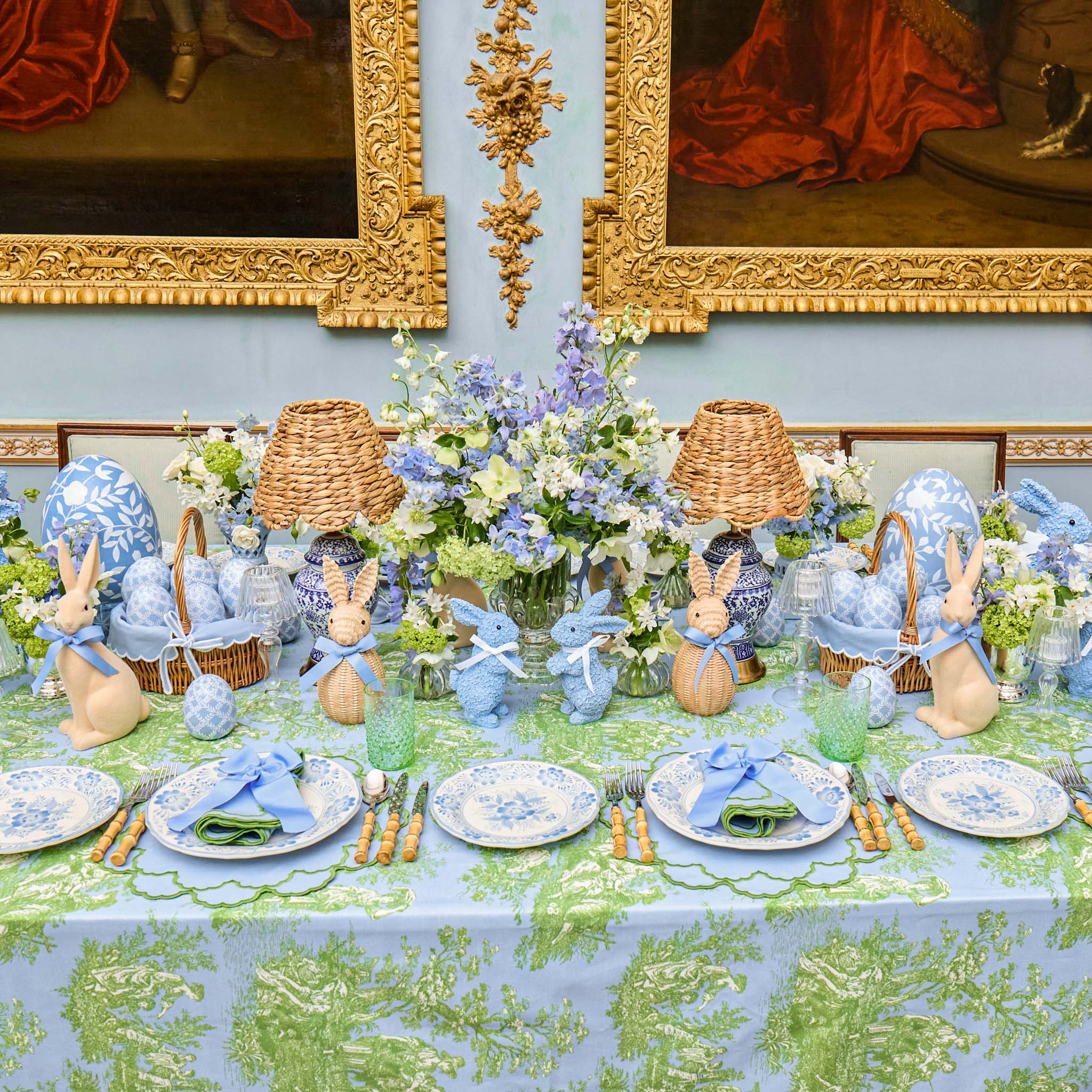 Easter table setting with decorative elements, including bunny figurines and floral arrangements, against a classical painting backdrop.