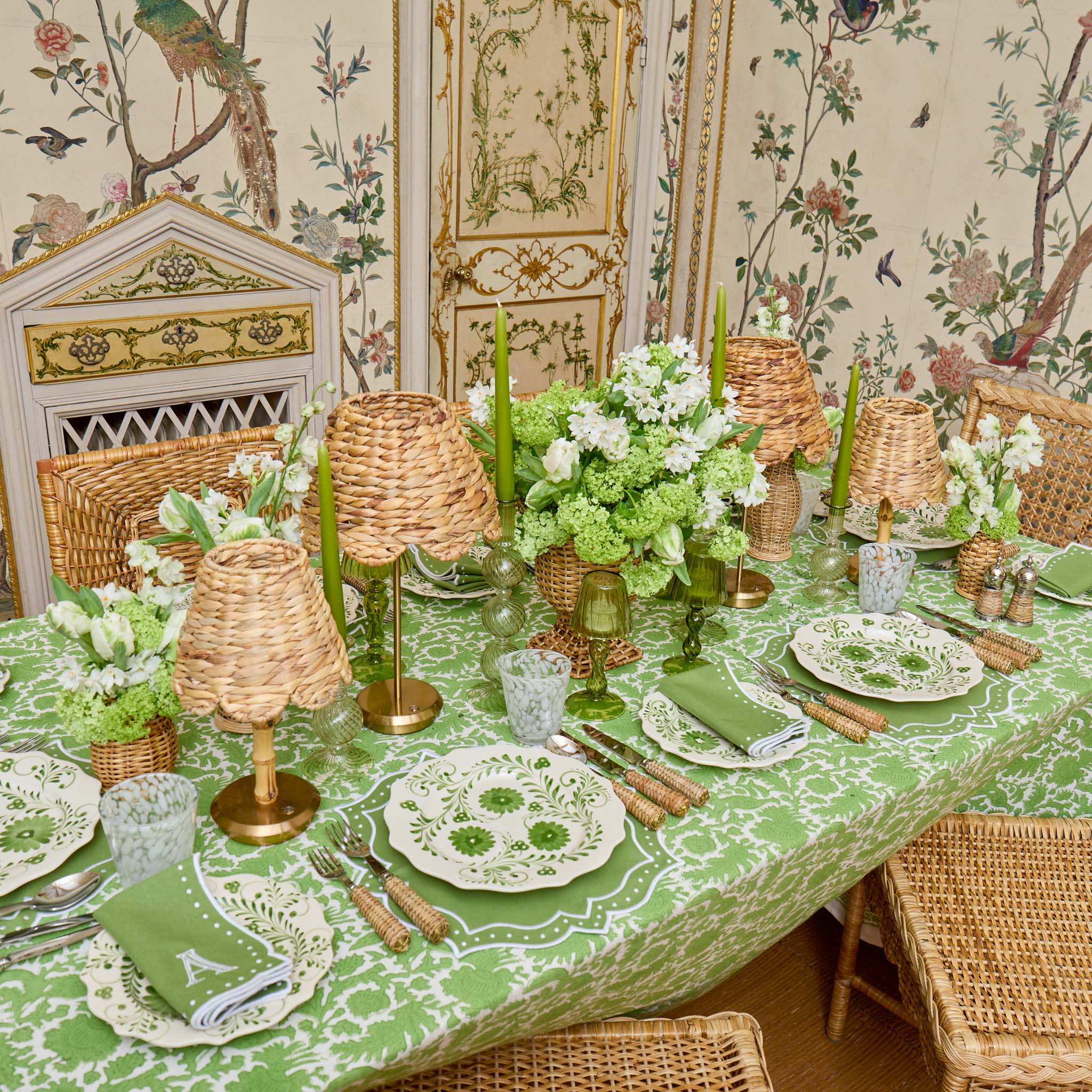 Elegant dining table setting with green tablecloth, floral centerpieces, and wicker chairs in a decorated room.