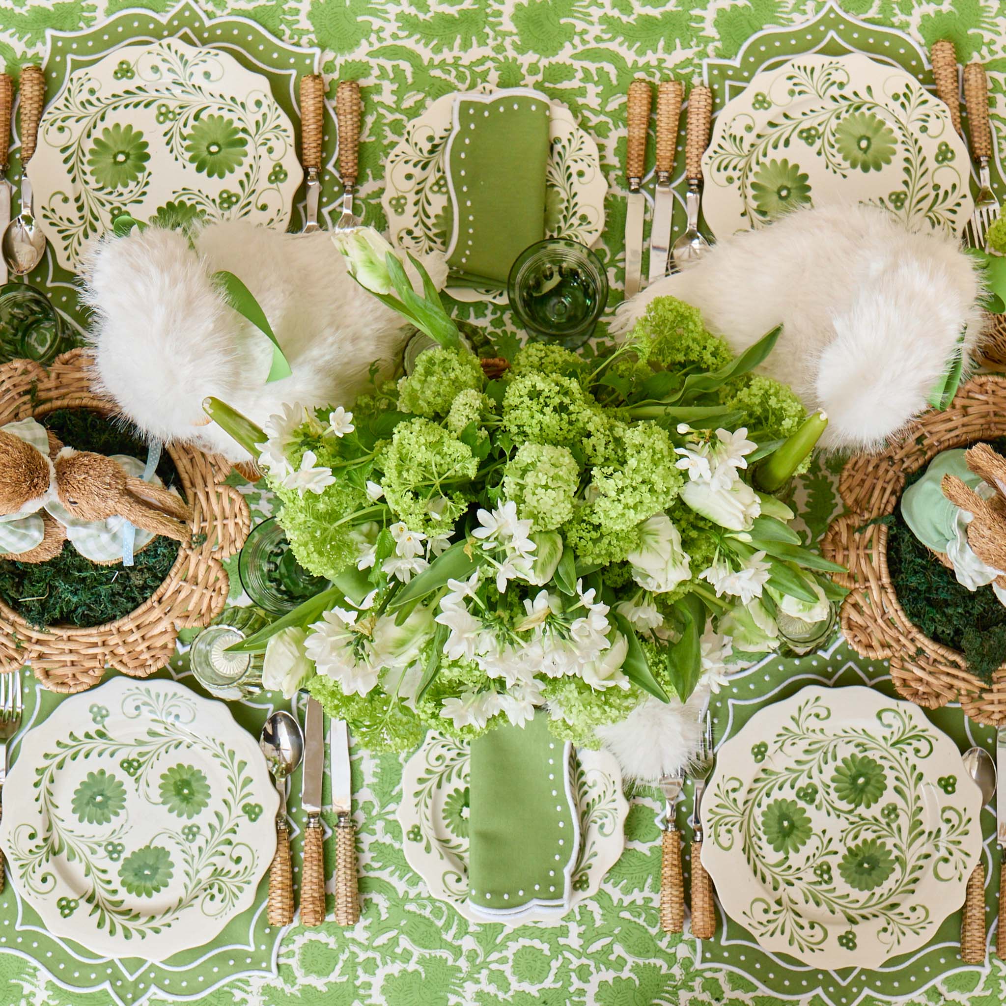 Decorative table setting with green and white floral centerpiece, patterned plates, and cutlery on a matching tablecloth.