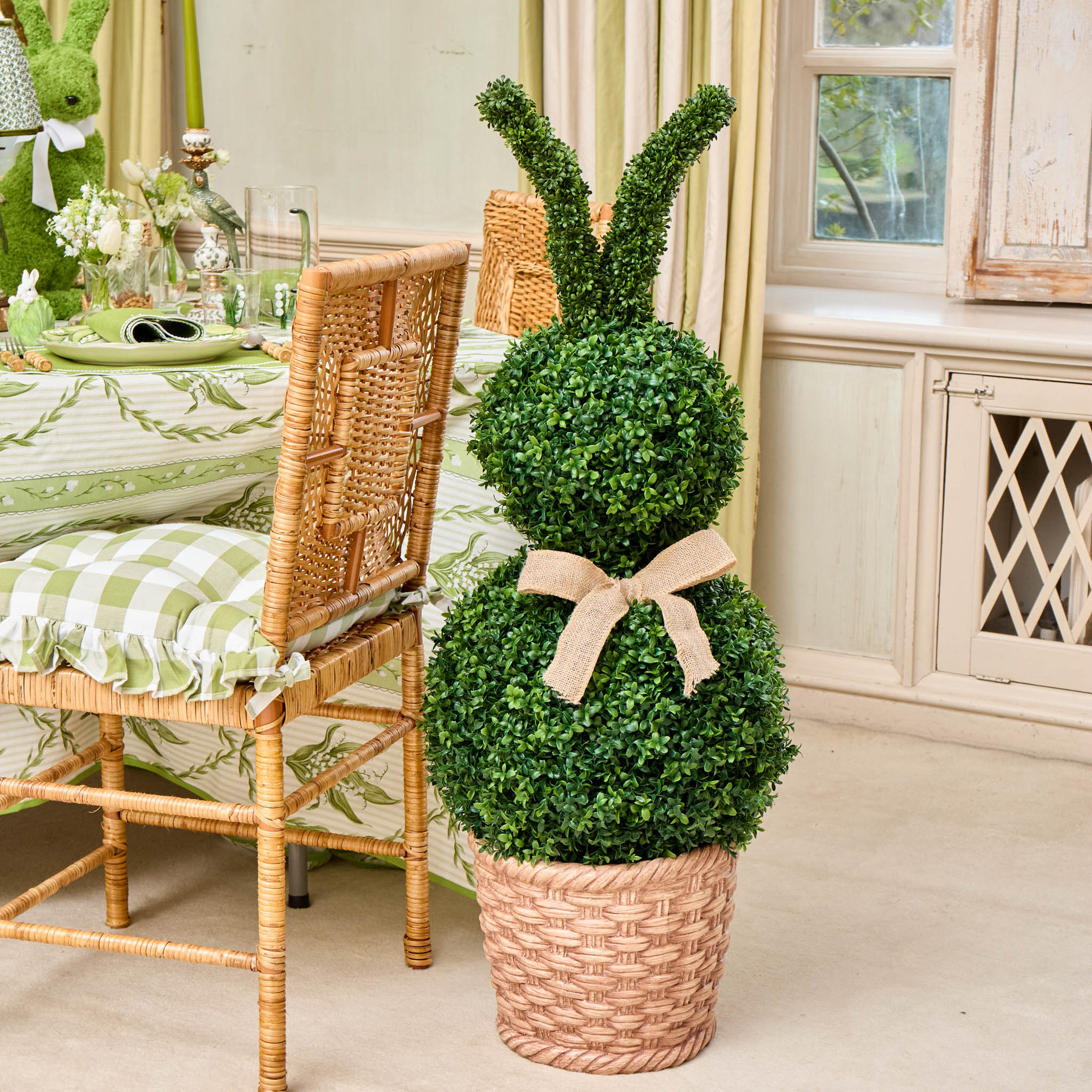 Decorative topiary in a woven basket with a bow, placed on a floor next to a table with a green and white checkered tablecloth.