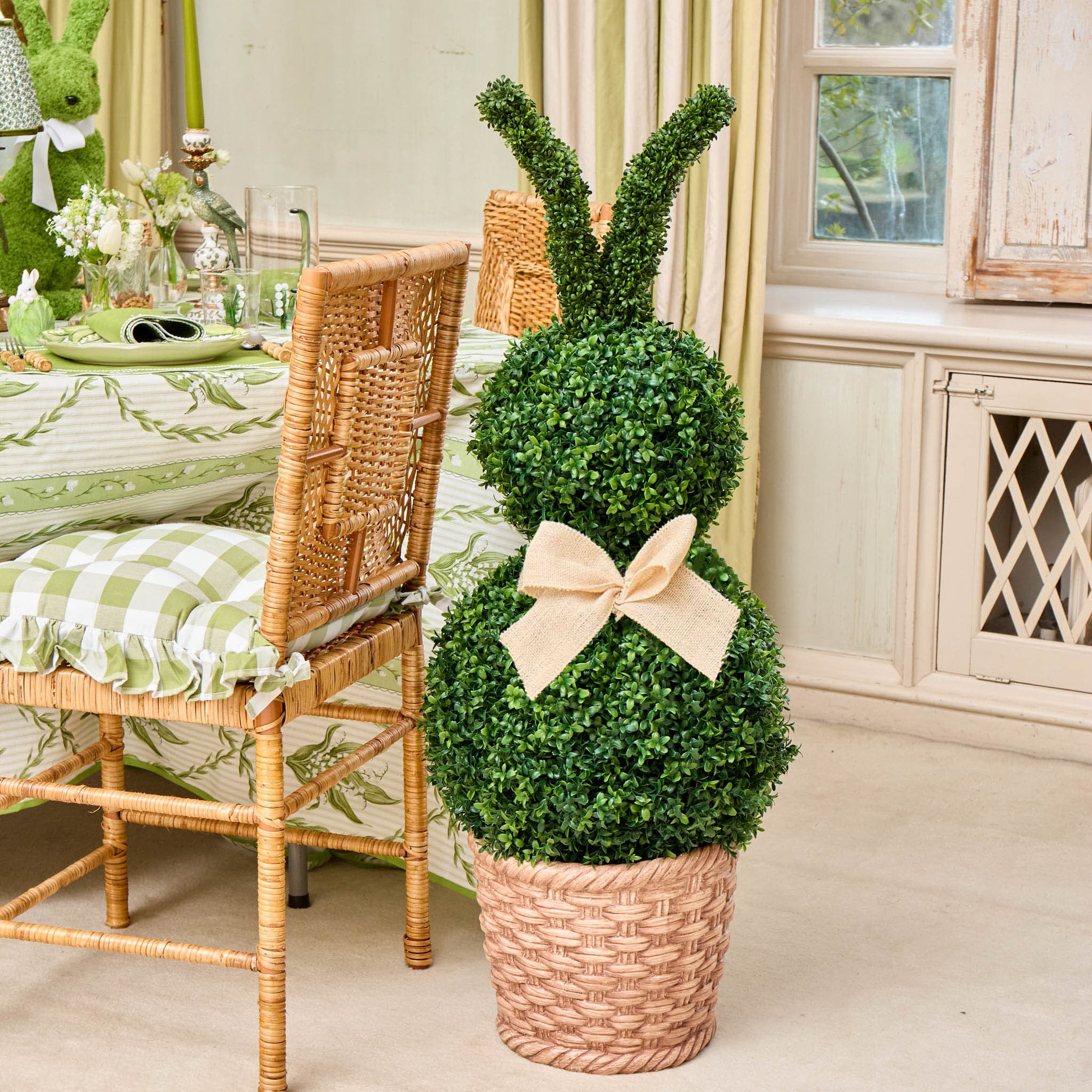Decorative topiary in a woven basket with a bow, placed on a floor next to a table with a green and white checkered tablecloth.