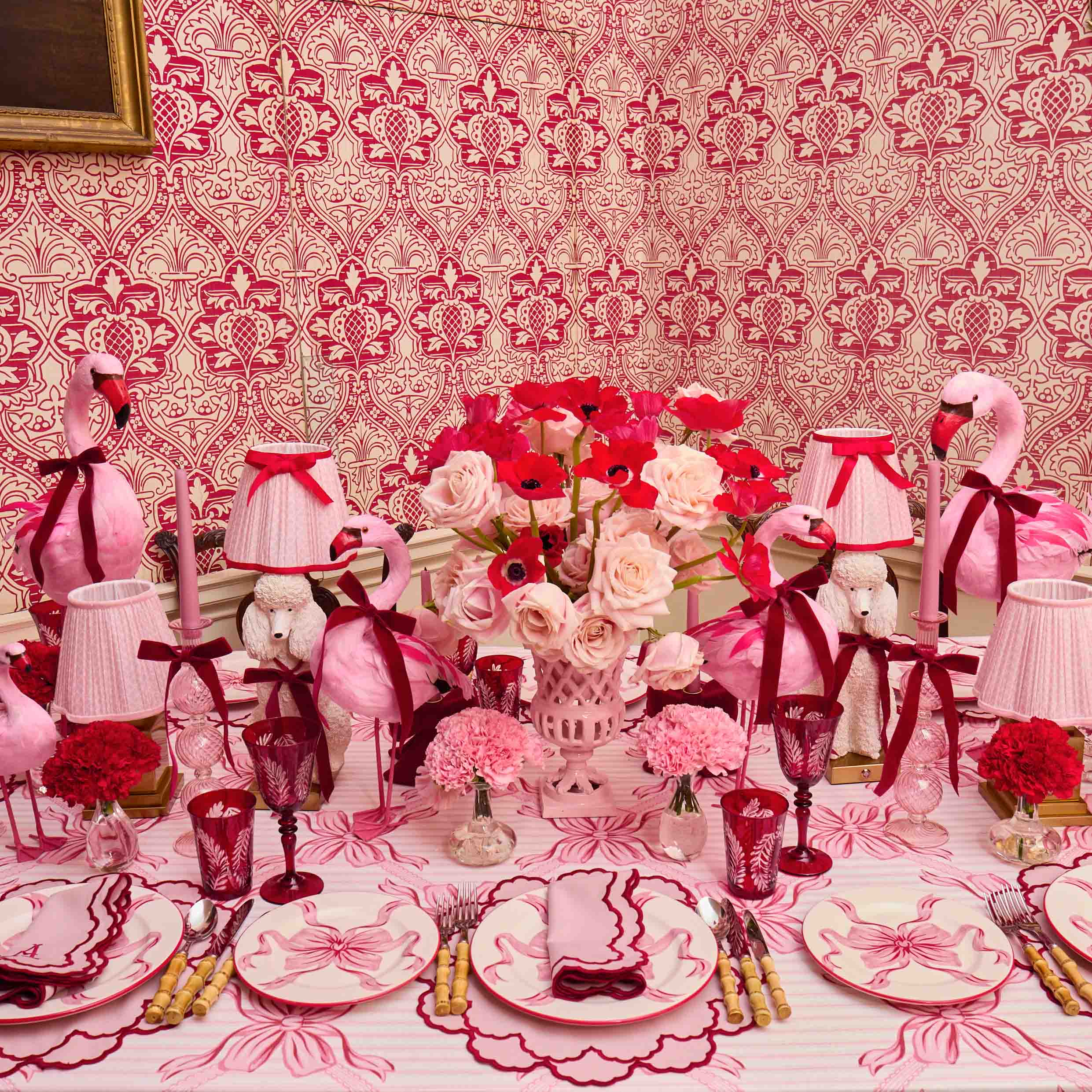 Decorative table setting with pink flamingo napkins, flowers, and tableware against a patterned red and white wall.
