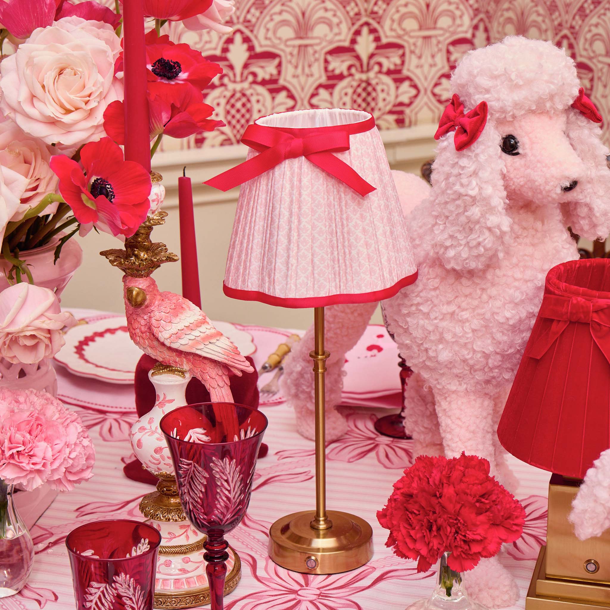 Decorative table setting with pink and red flowers, a lamp, and a plush toy on a patterned wallpaper background.