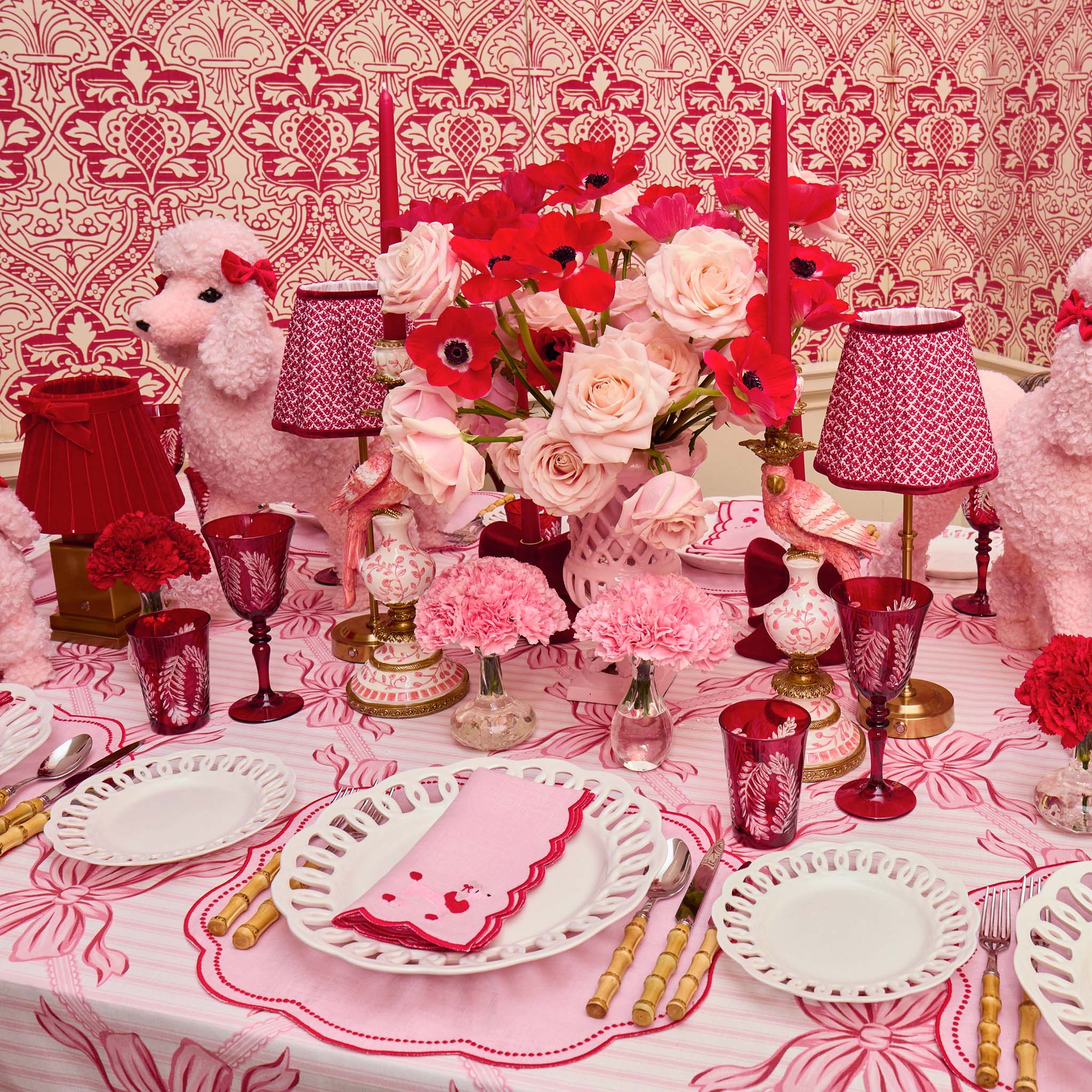 Decorative table setting with pink and red flowers, lamps, and tableware against a patterned wallpaper.