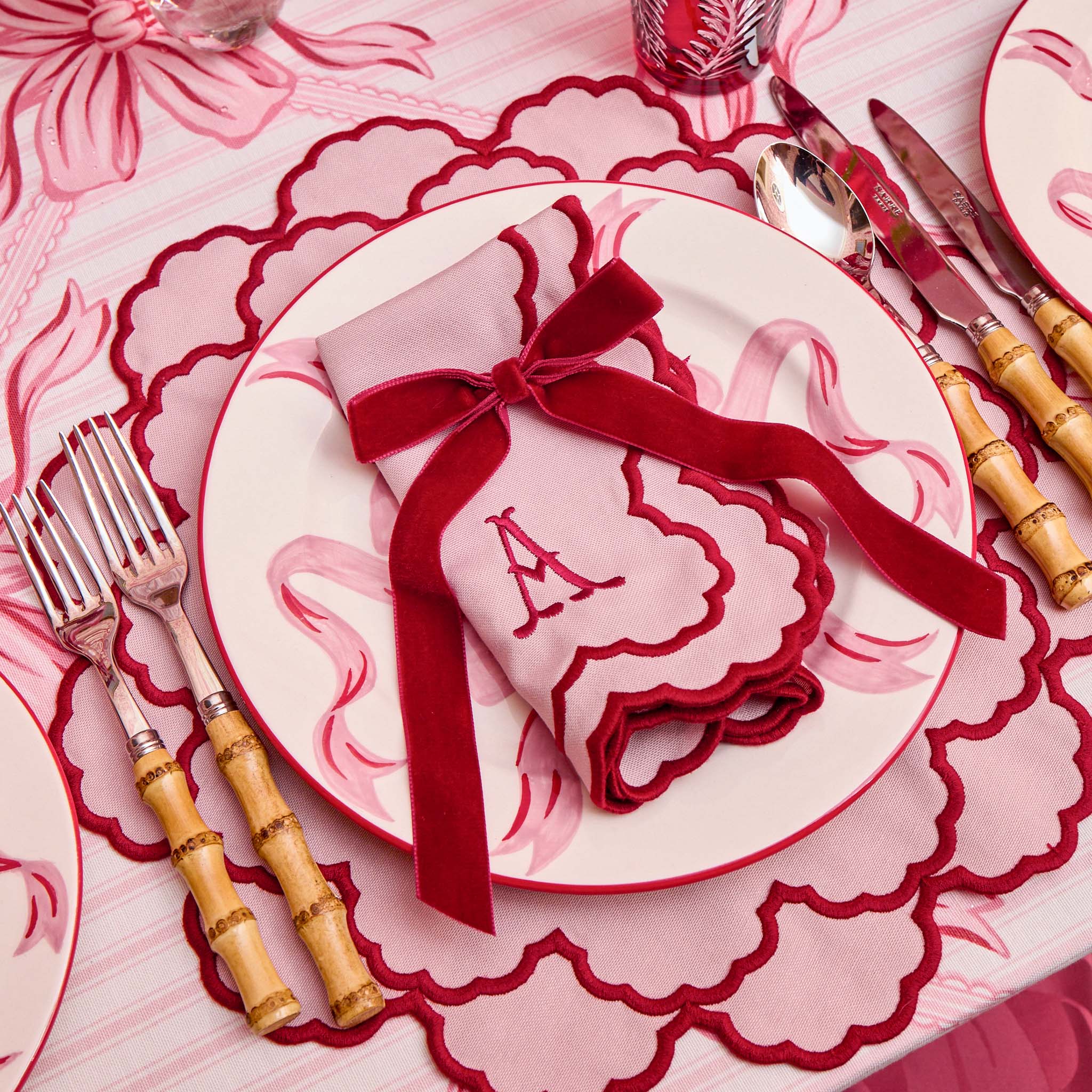 Dining table setting with a red napkin tied with a bow on a white plate, surrounded by cutlery and a floral tablecloth.