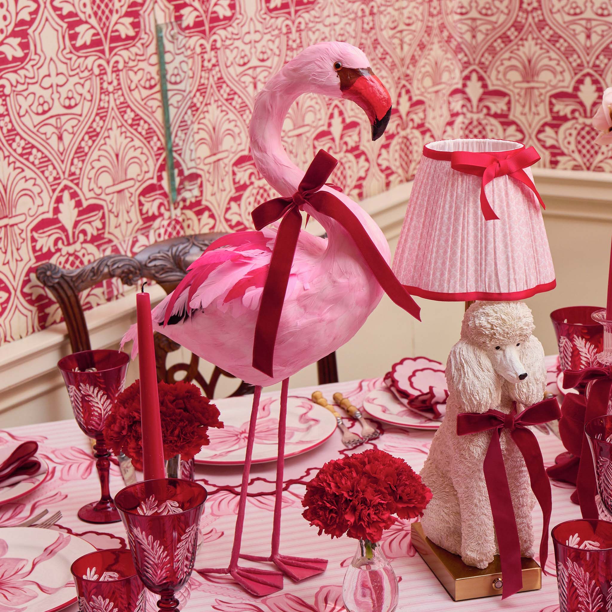 Decorative table setting with a pink flamingo, poodle lamp, and red accents against a patterned wall.