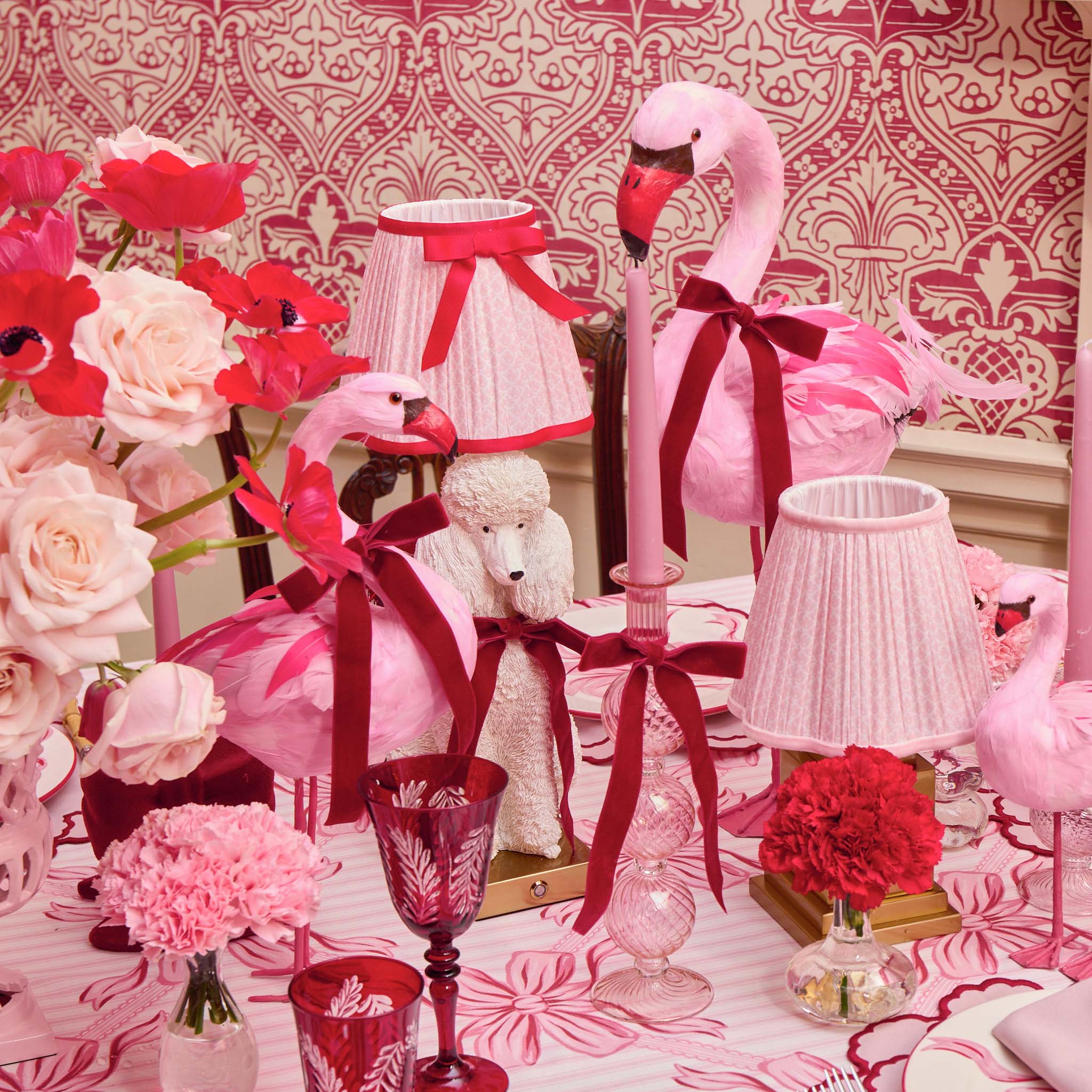 Decorative table setting with pink flamingo figurines, floral arrangements, and lamps against a patterned wall.