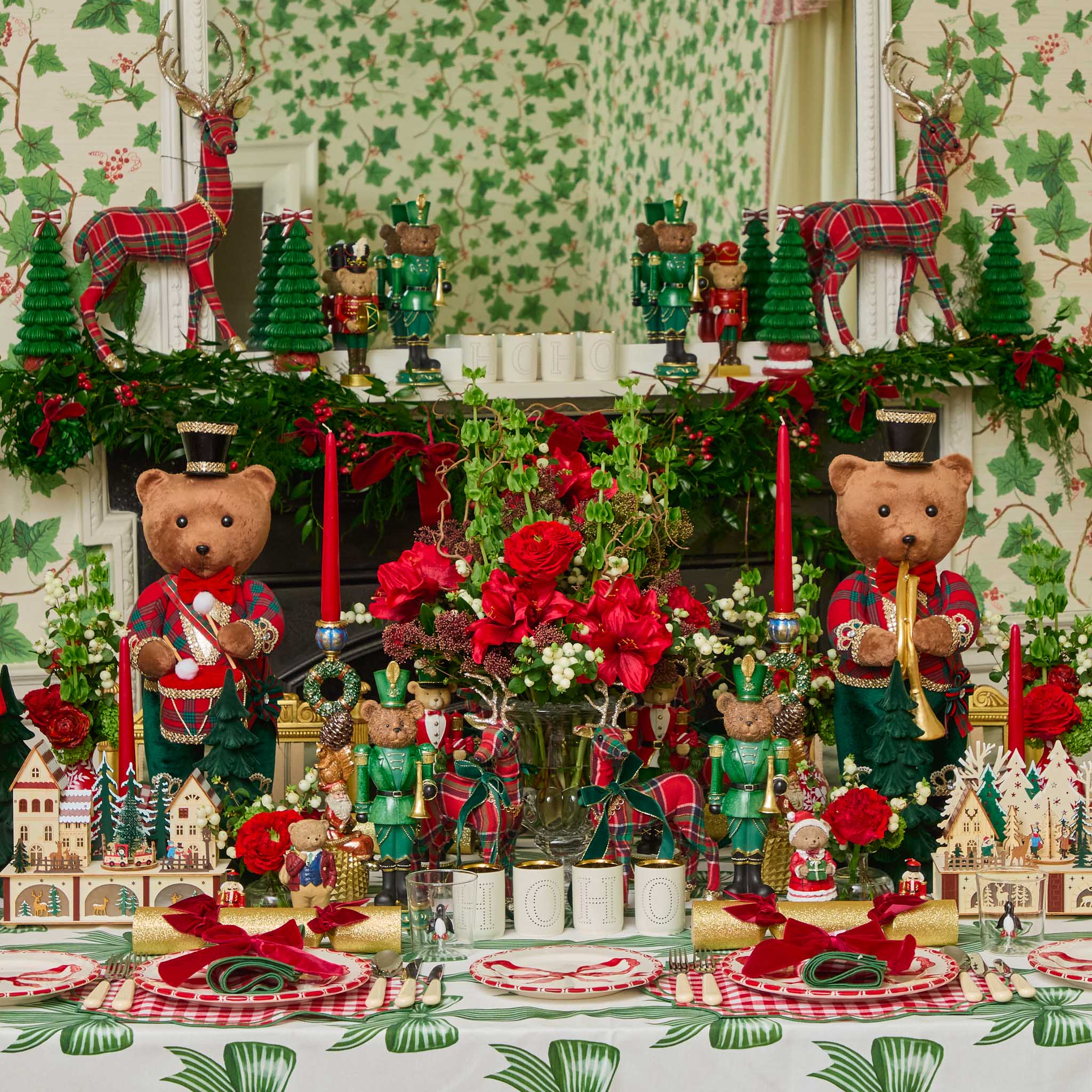 Decorative Christmas table setting with teddy bears, candles, and greenery against a patterned wallpaper.
