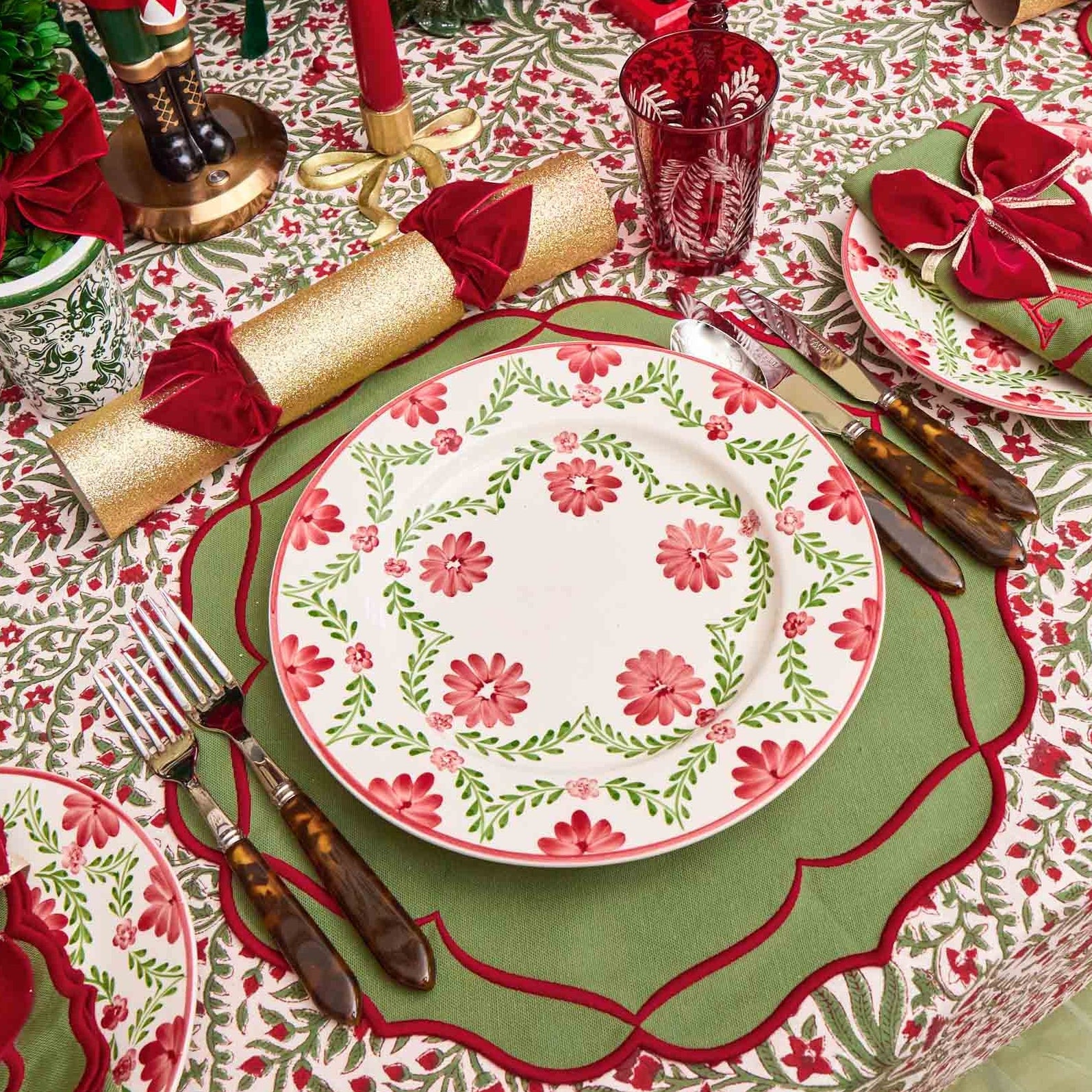 Christmas-themed table setting with floral plates, red napkins, and decorative cutlery on a patterned tablecloth.