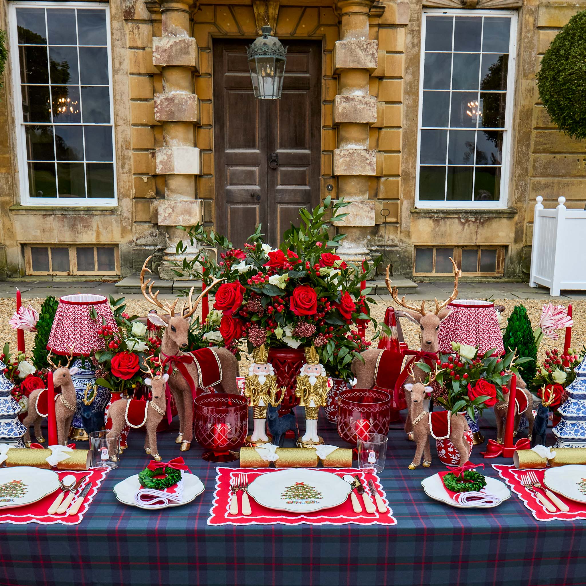 Decorative Christmas table setting with reindeer and red tableware in front of a stone building.