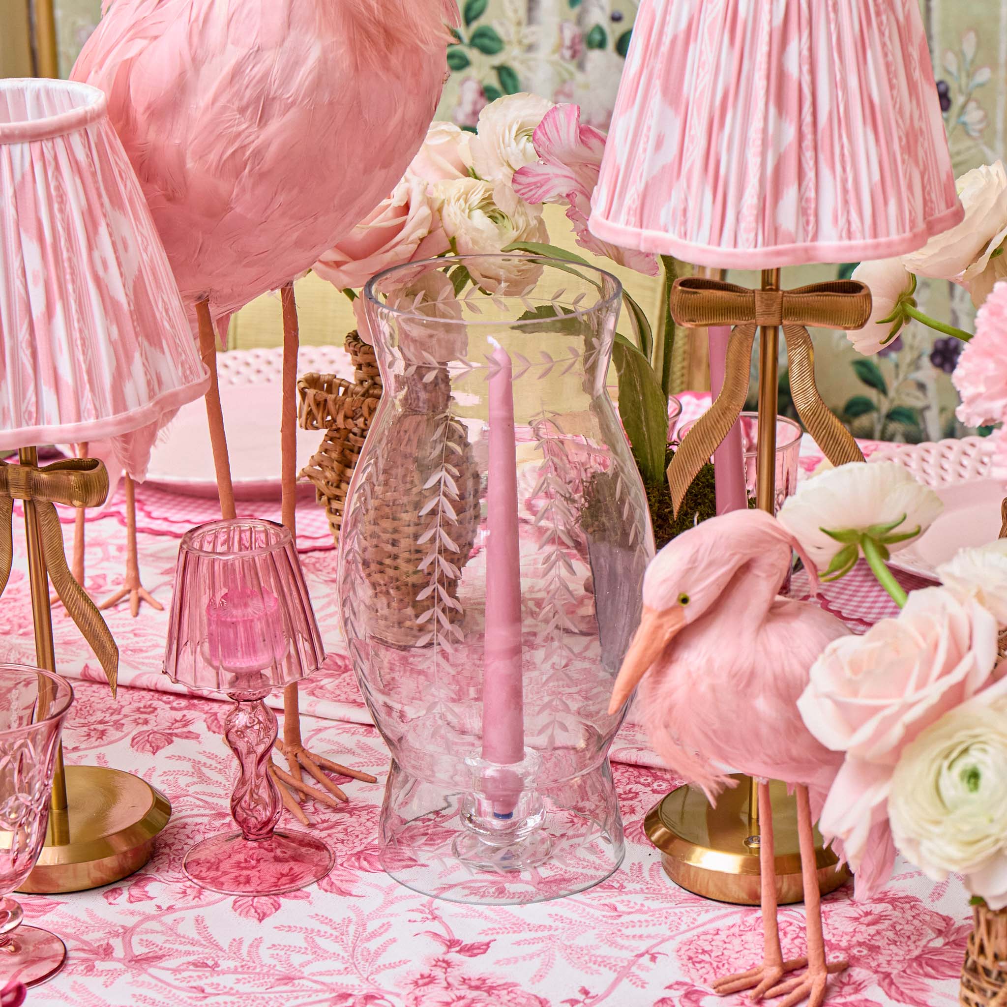 Decorative setting with pink flamingo figurines, a glass vase, and pink lamps on a floral tablecloth.