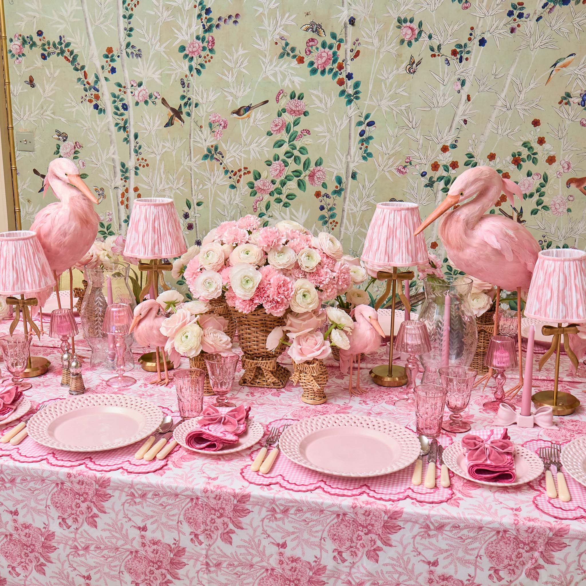 Pink flamingo-themed table setting with floral arrangements and decorative lamps against a floral wallpaper background.