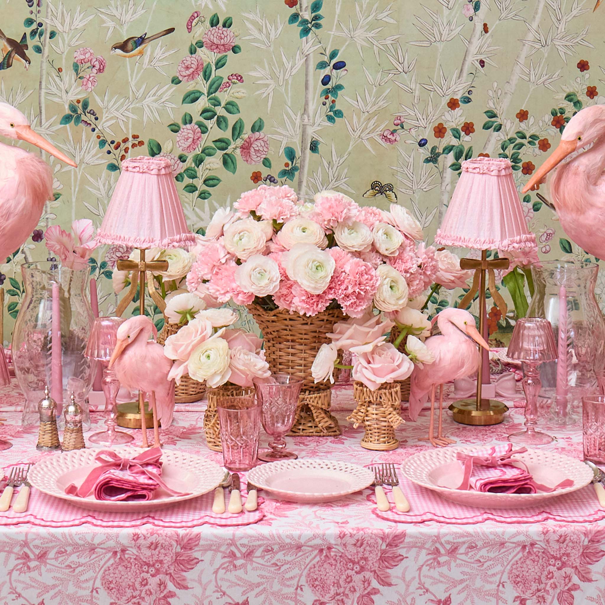 Pink flamingo-themed table setting with floral arrangements and lamps against a floral wallpaper background.