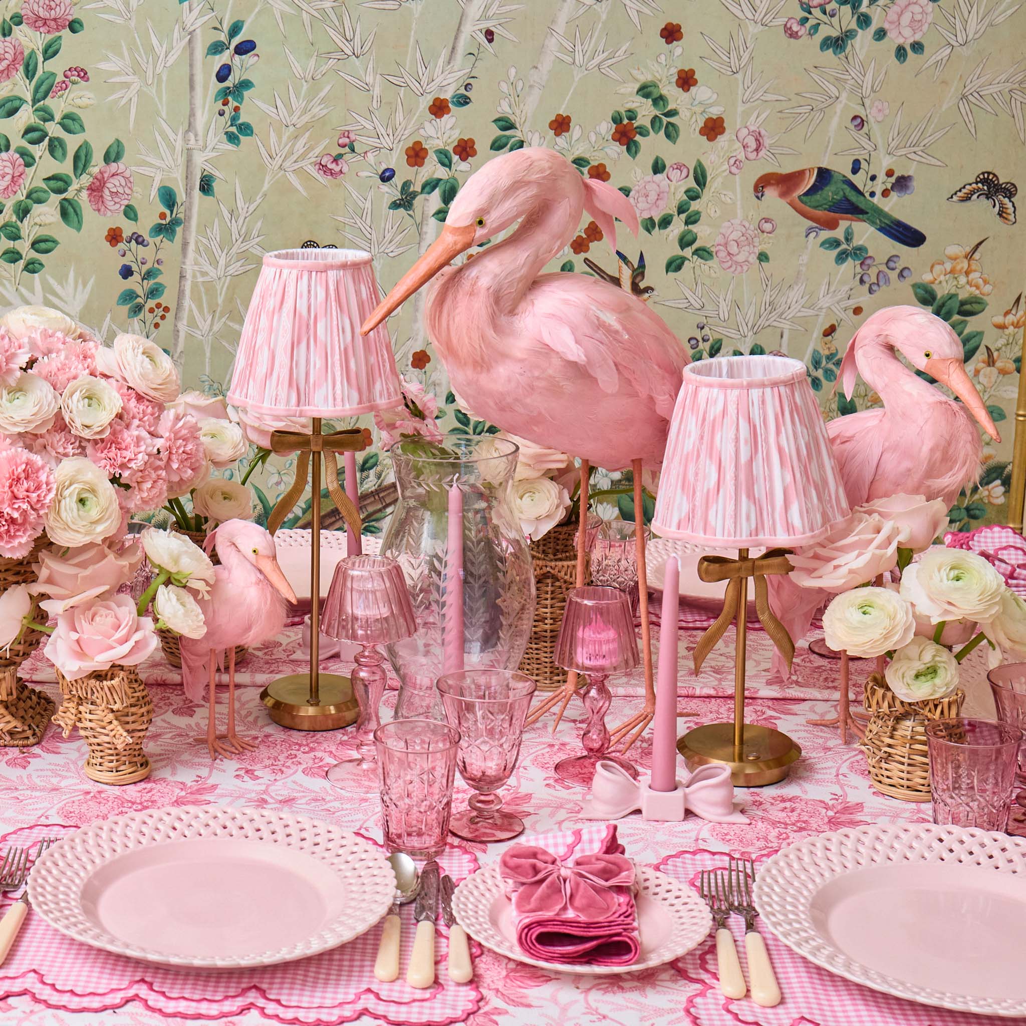 Pink flamingo decor on a table with floral arrangements and lamps against a floral wallpaper background.
