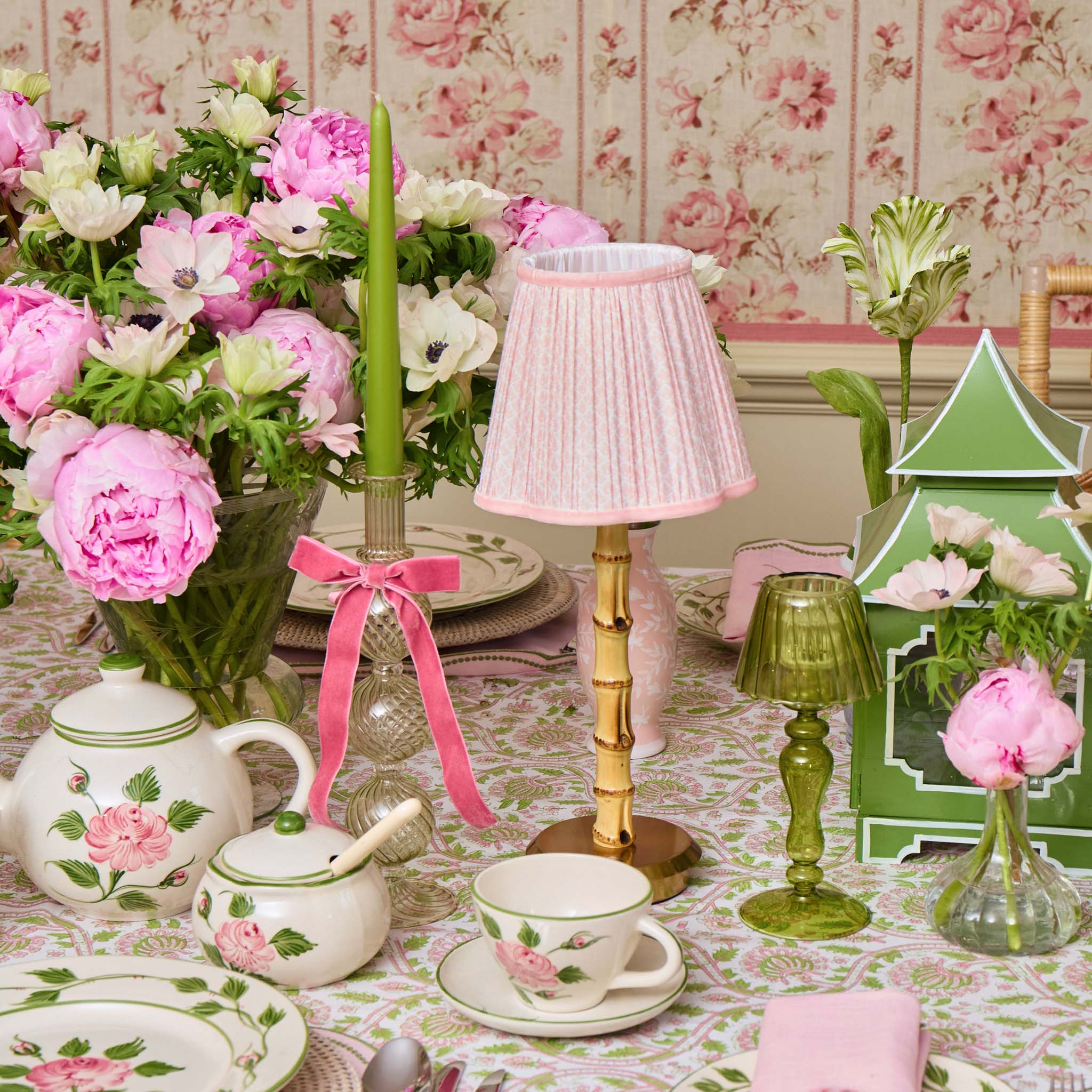 Decorative table setting with floral teacup, teapot, and table lamp against a floral wallpapered wall.