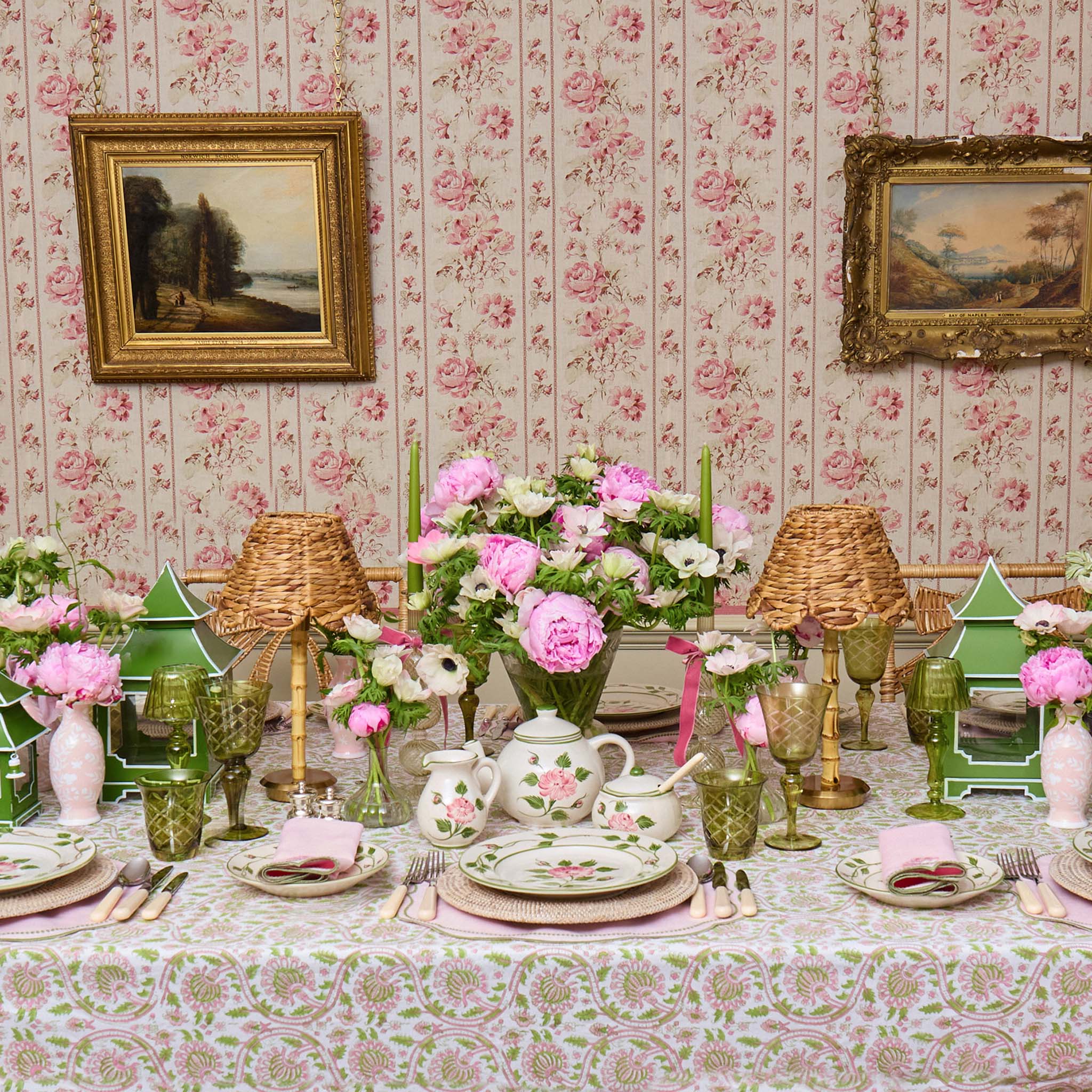 Decorative table setting with floral tablecloth, teapot, and candles in a room with floral wallpaper.