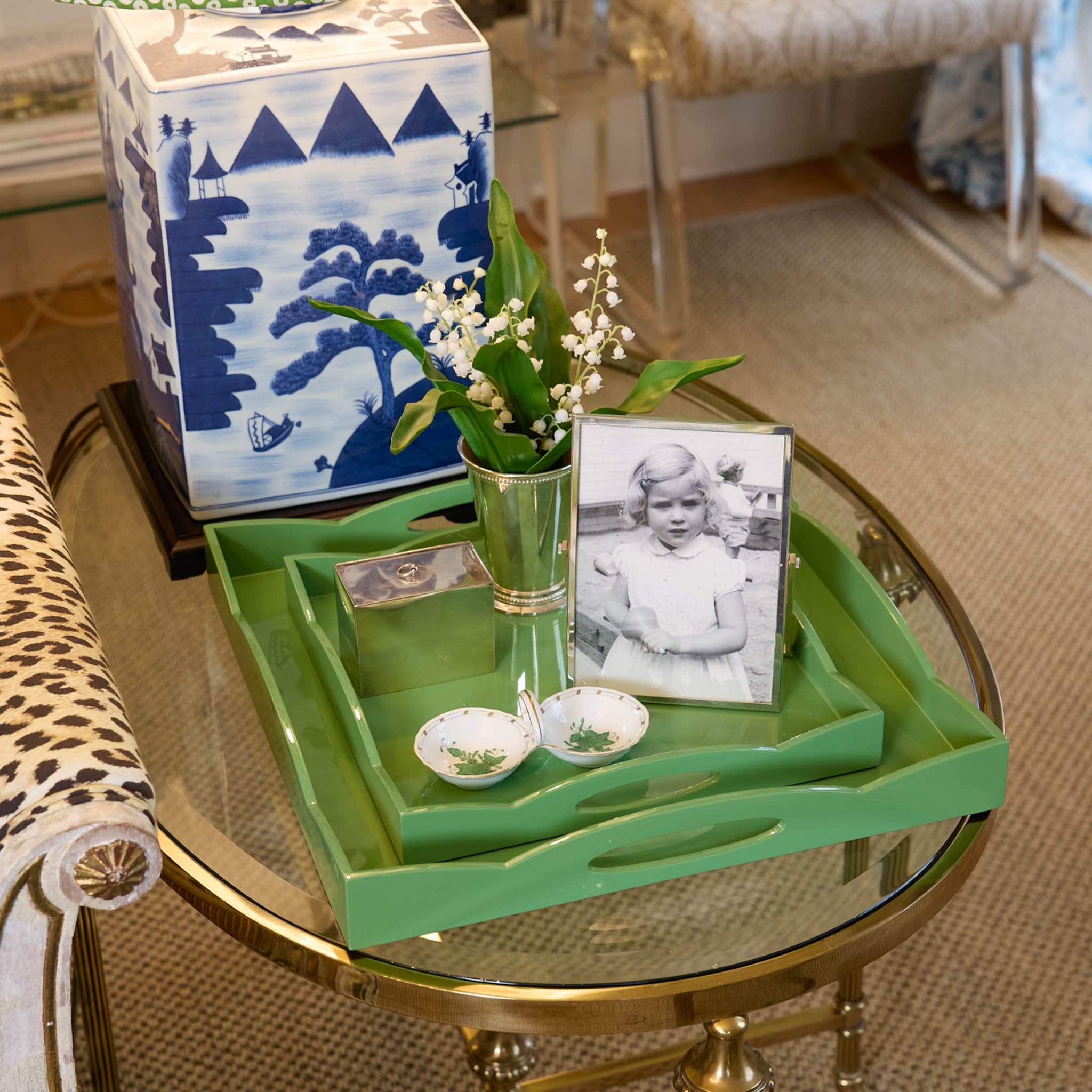 Green tray with decorative items including a vase with flowers, small photo frame, and blue and white ceramic box on a glass table.