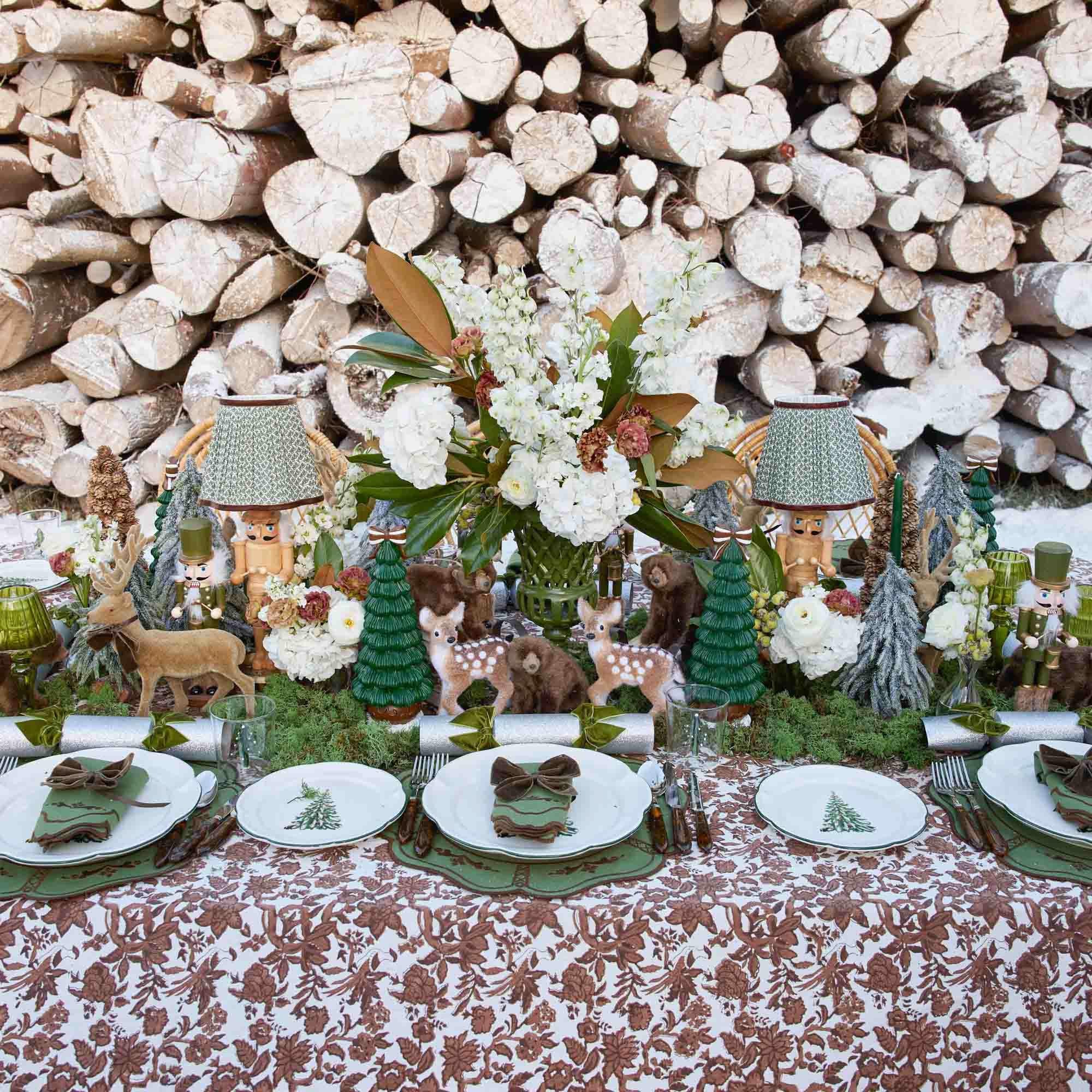 Wooden log pile with a festive table setting featuring deer, trees, and floral arrangements.