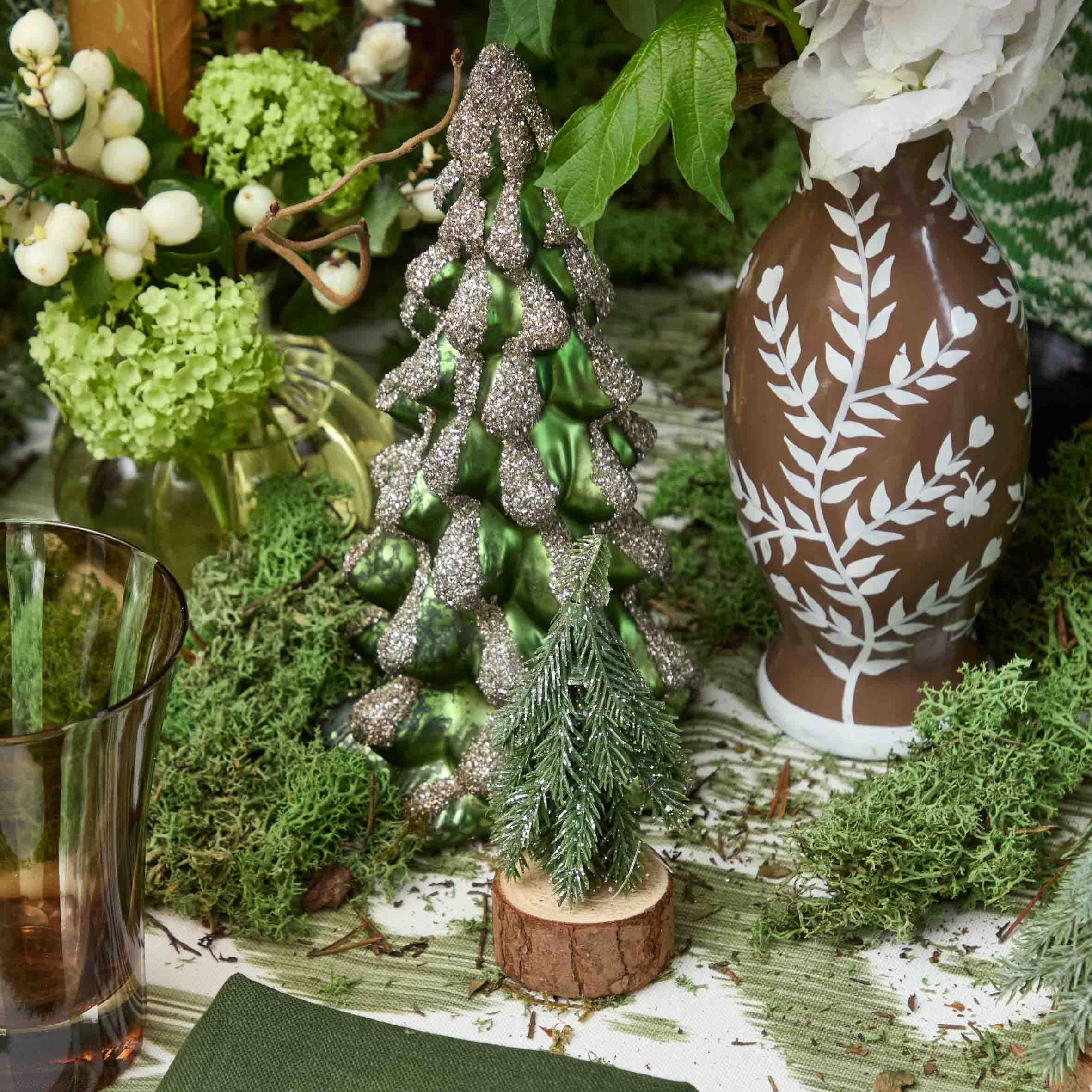 Decorative Christmas tree with glittery green and silver finish on a wooden base, surrounded by greenery and a patterned vase.
