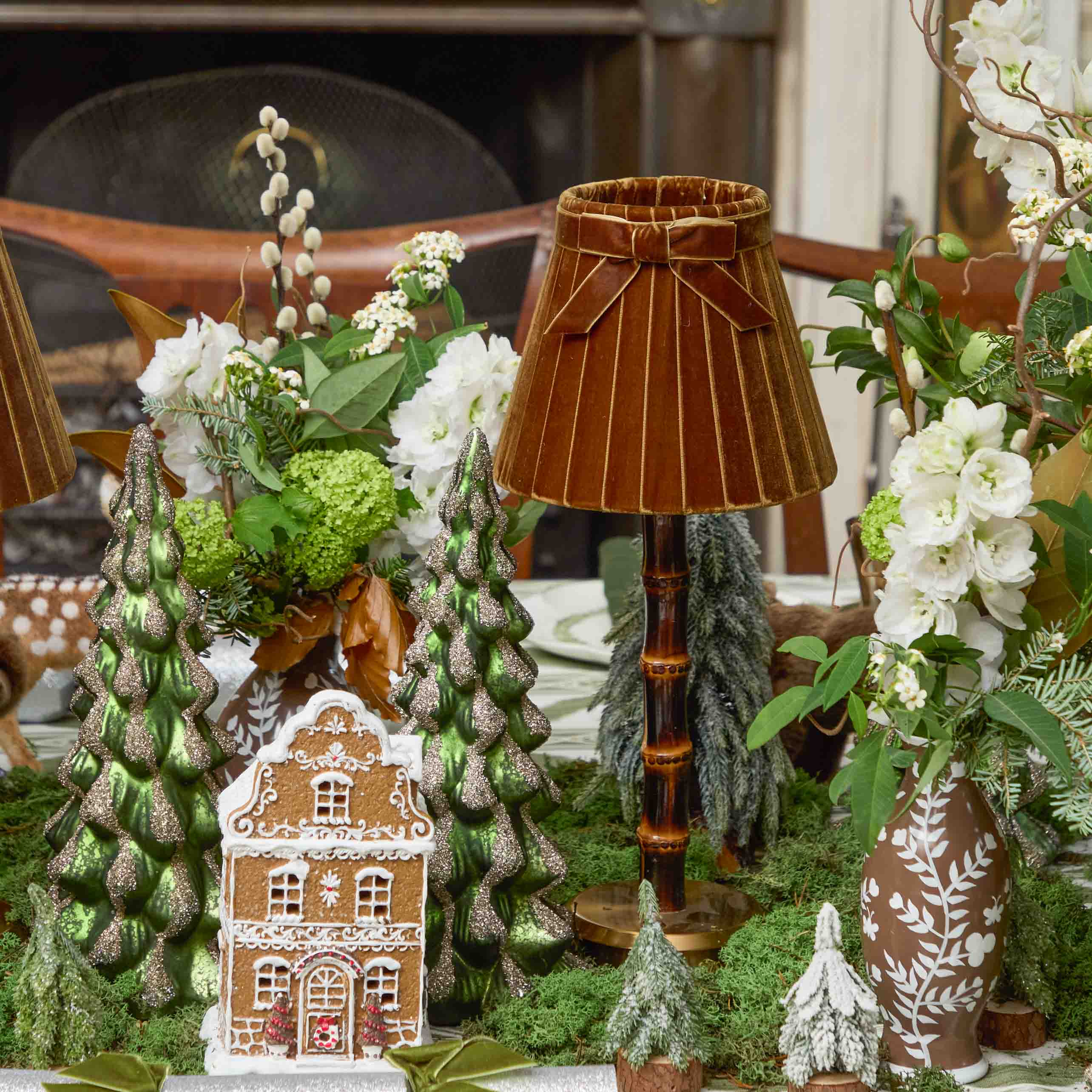 Decorative table setting with gingerbread house, Christmas trees, and a lamp.