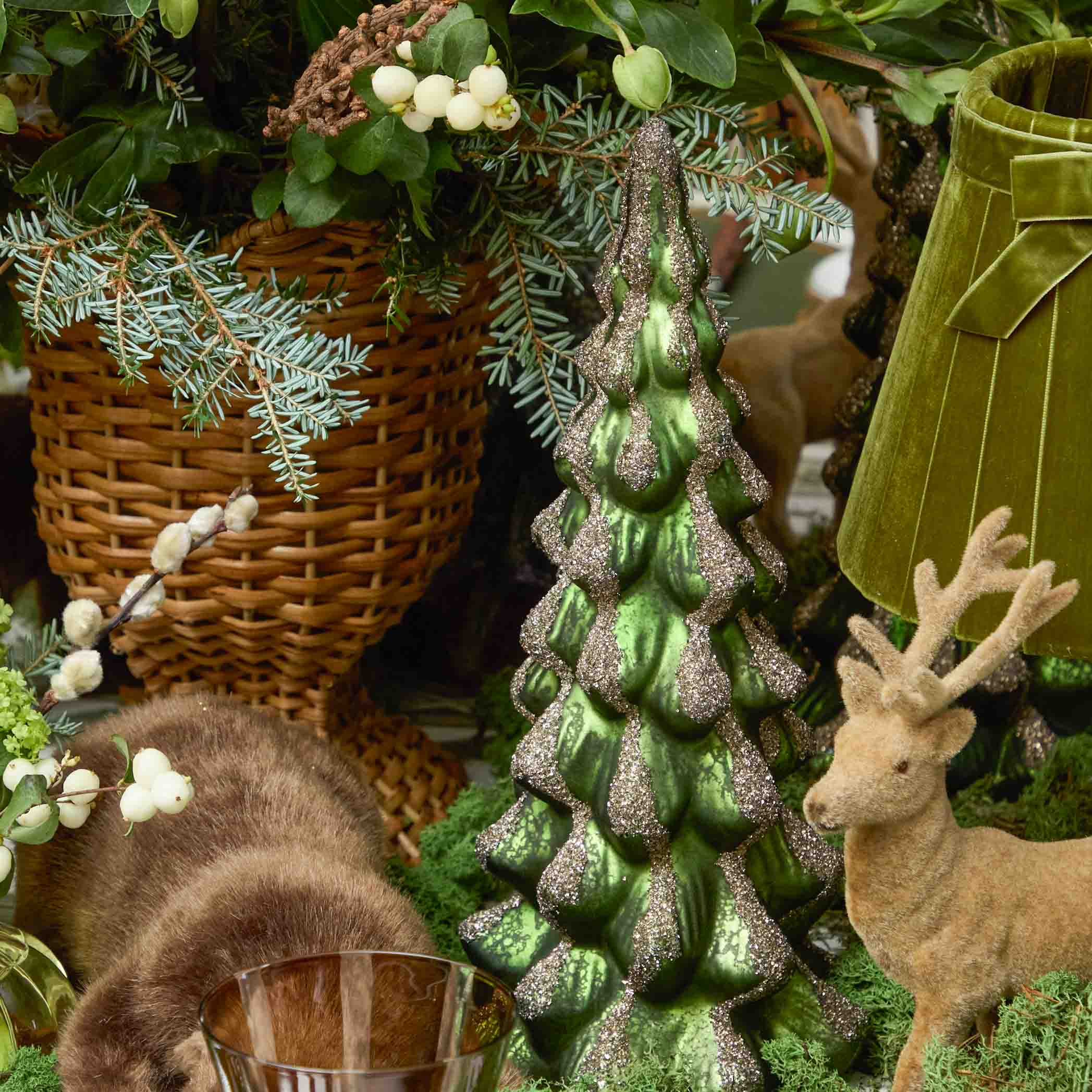Decorative scene with a green glittery tree, wicker basket, and deer figurine.