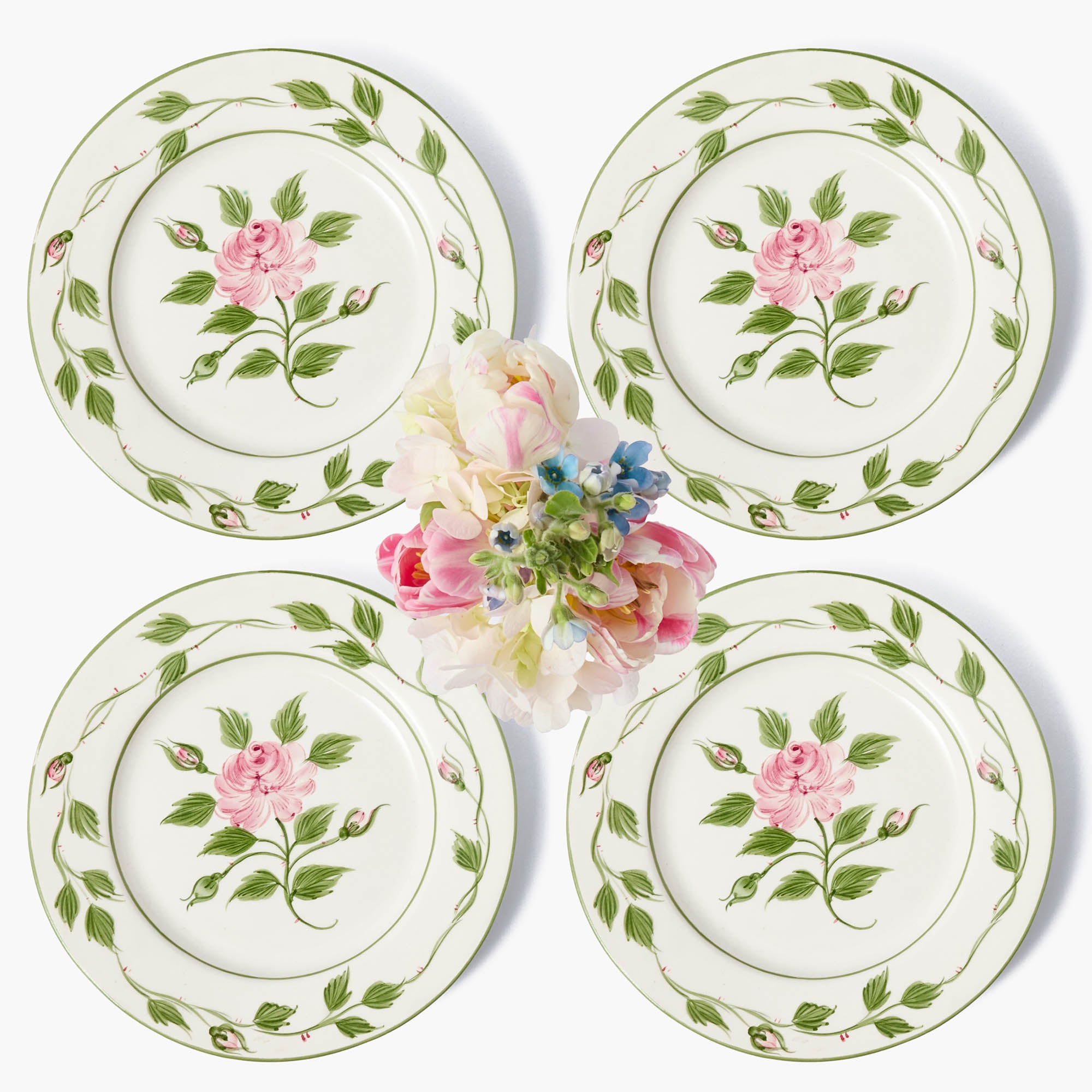A set of four white ceramic dinner plates with a green floral pattern and a rose motif in the center, displayed with a bouquet of flowers.