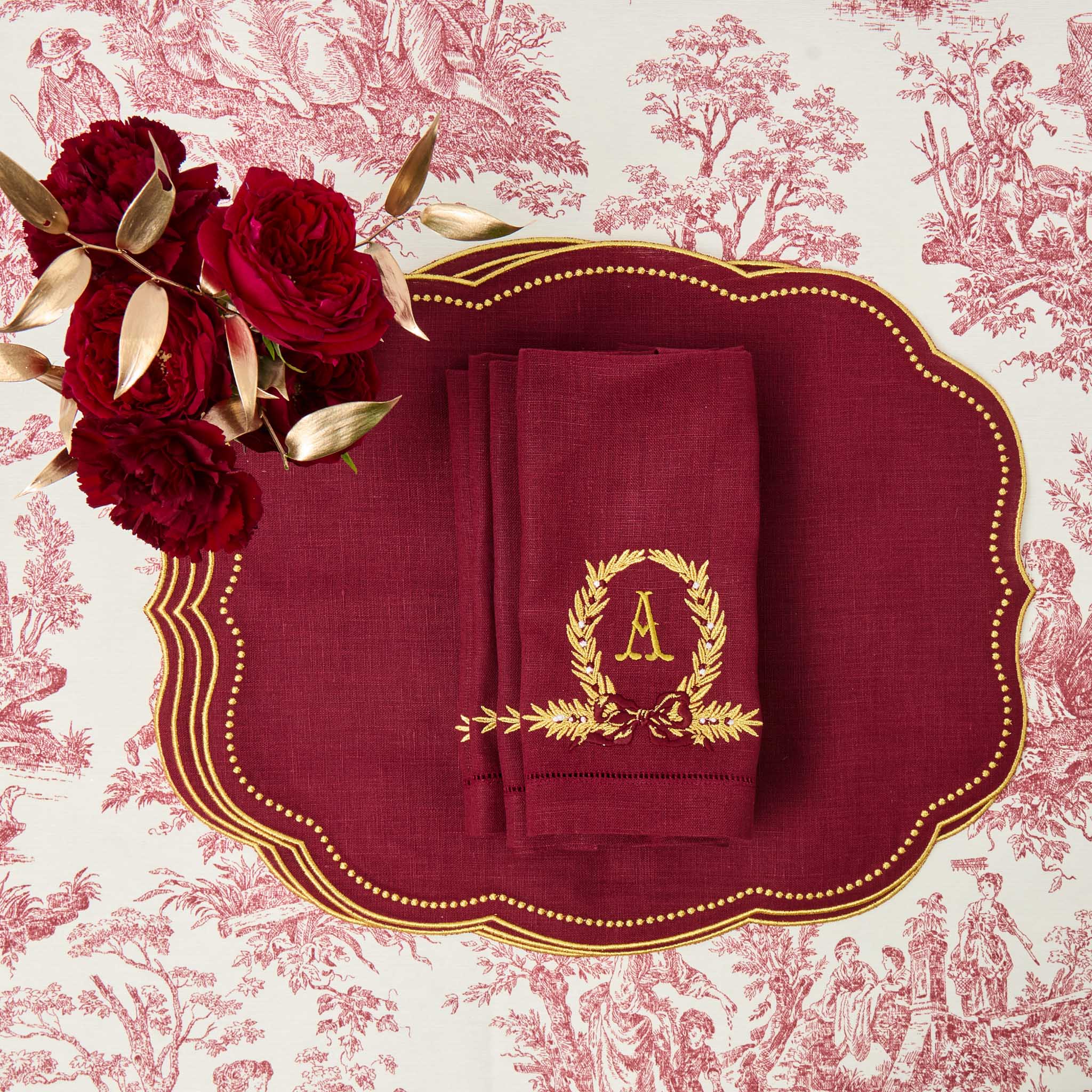 Maroon linen napkin with gold monogram on a decorative plate with red roses against a toile background