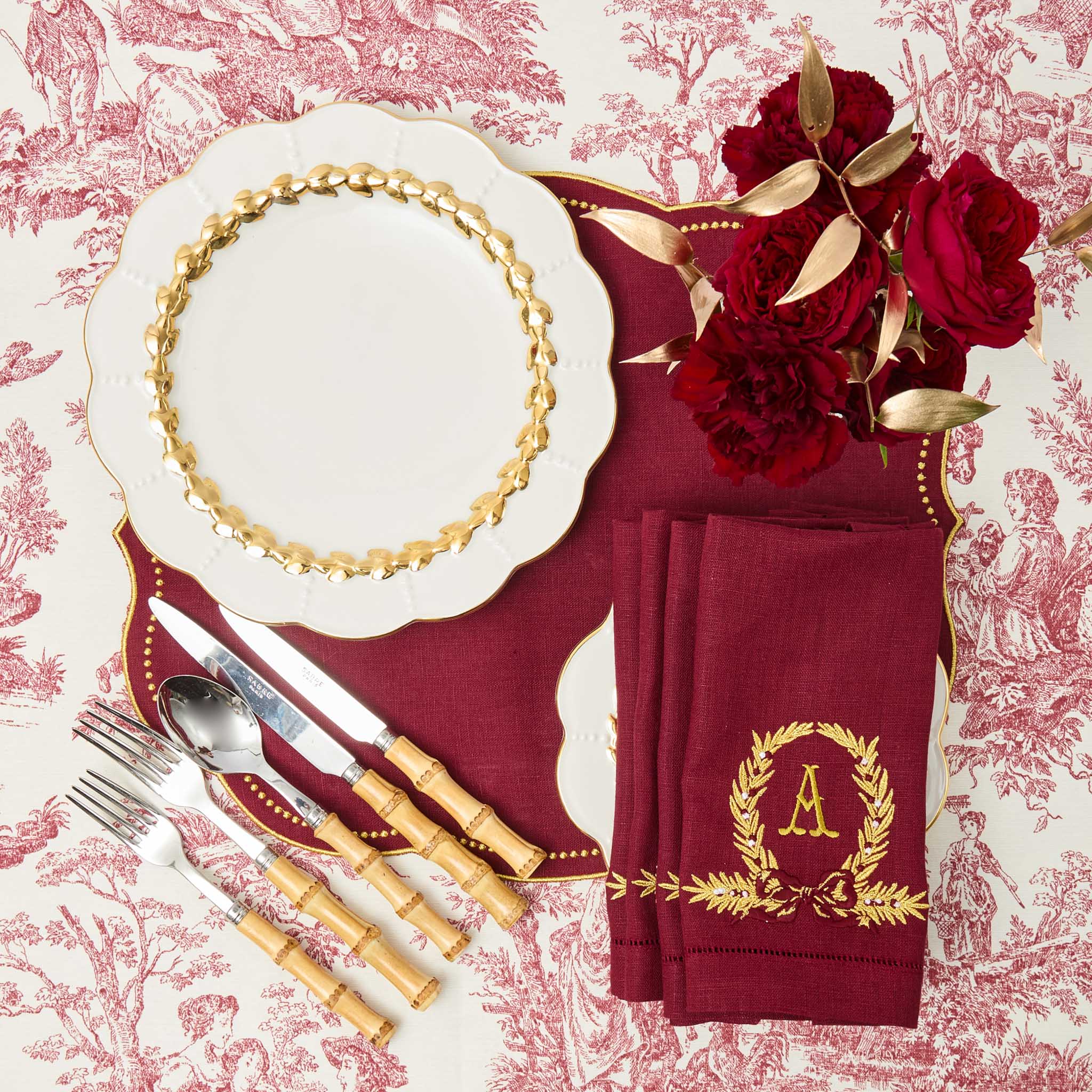 Elegant table setting with red napkins, gold cutlery, and floral arrangement on a patterned tablecloth.