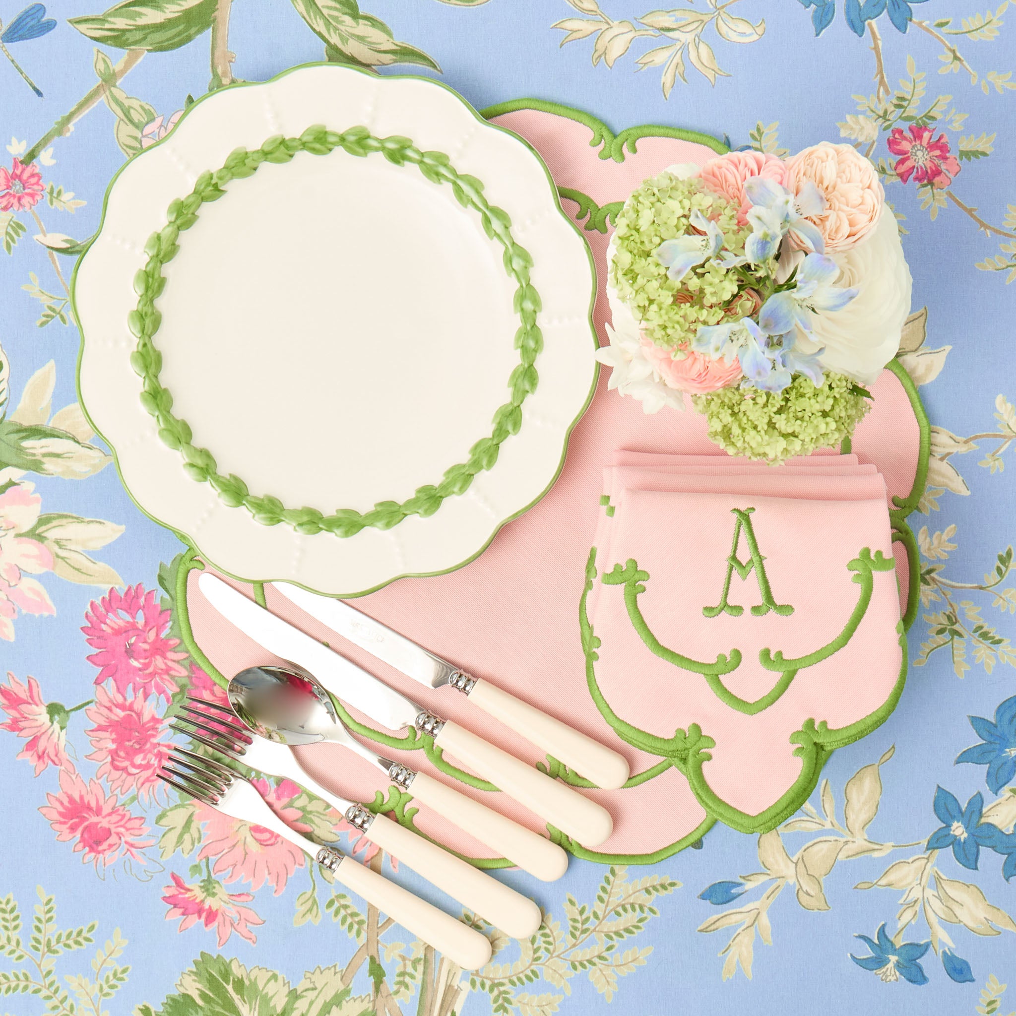 Vintage-style table setting with pink cutlery, floral centerpiece, and decorative plates on a floral-patterned background.