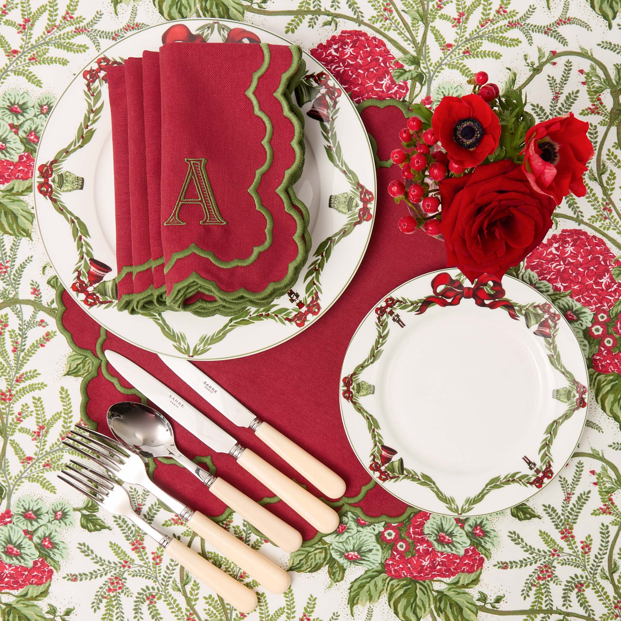 Christmas-themed table setting with red and green napkins, cutlery, and floral decorations on a patterned tablecloth.