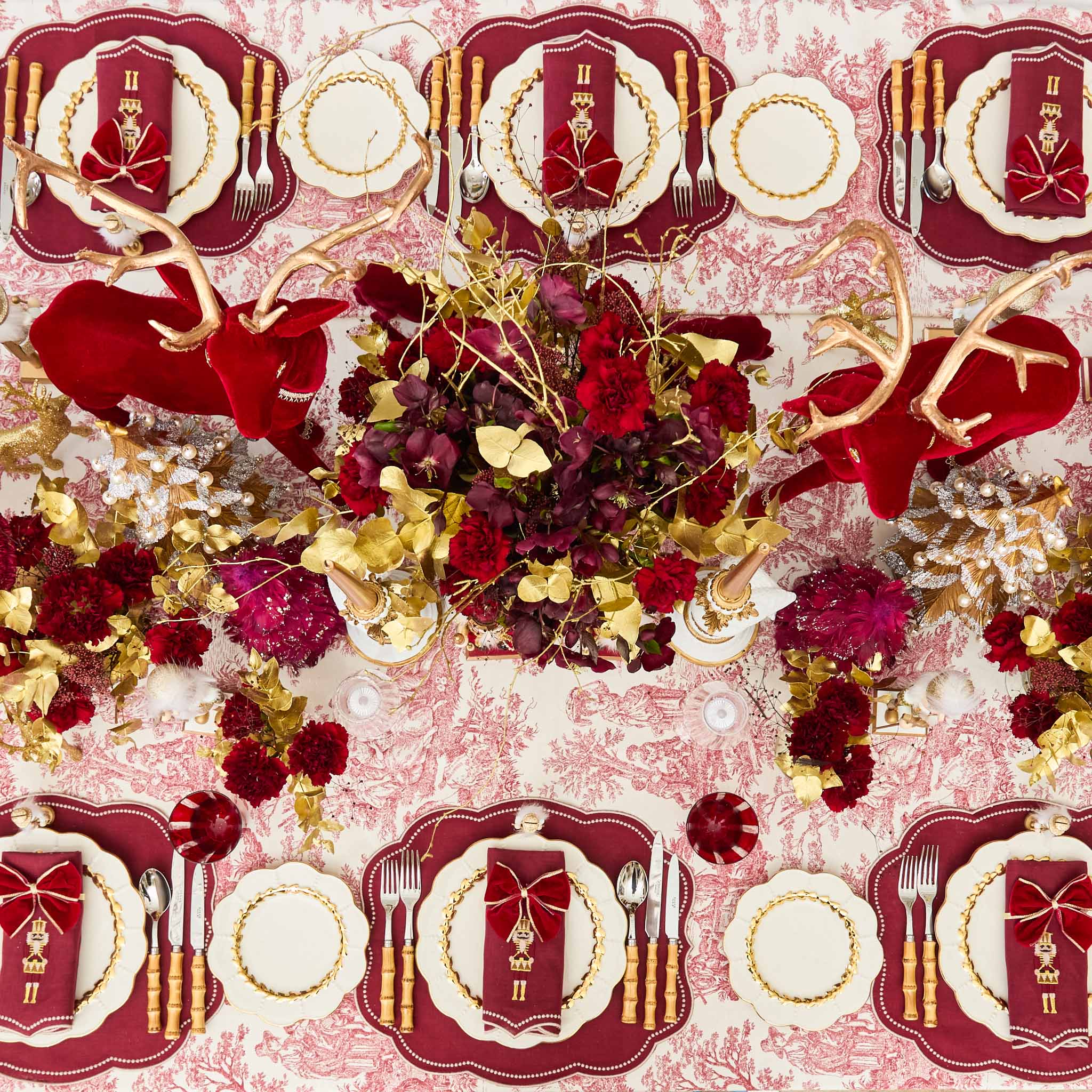 Decorative table setting with red and gold accents, flowers, and place cards on a pink floral patterned tablecloth.
