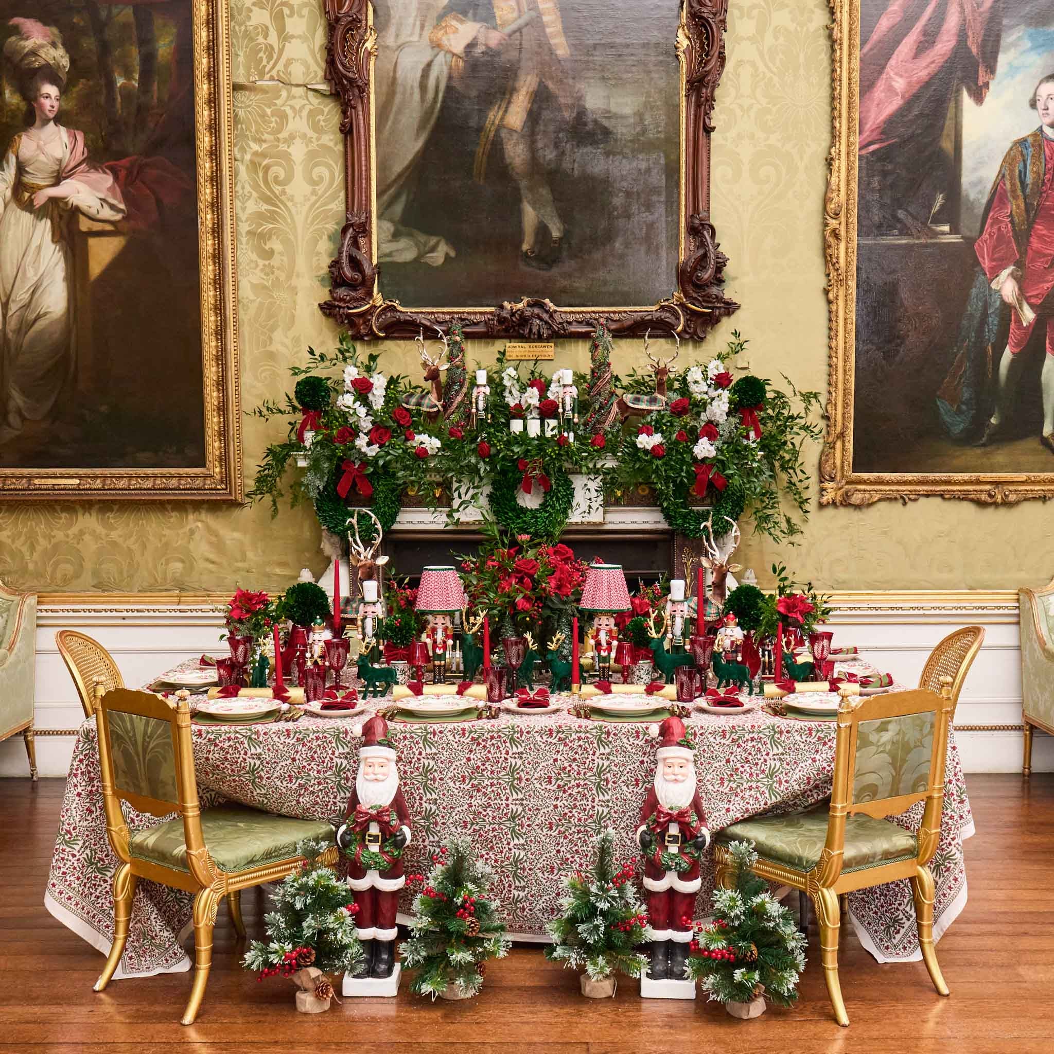 Decorated dining table with Christmas-themed centerpieces and Santa figurines, surrounded by classical paintings on a wall.