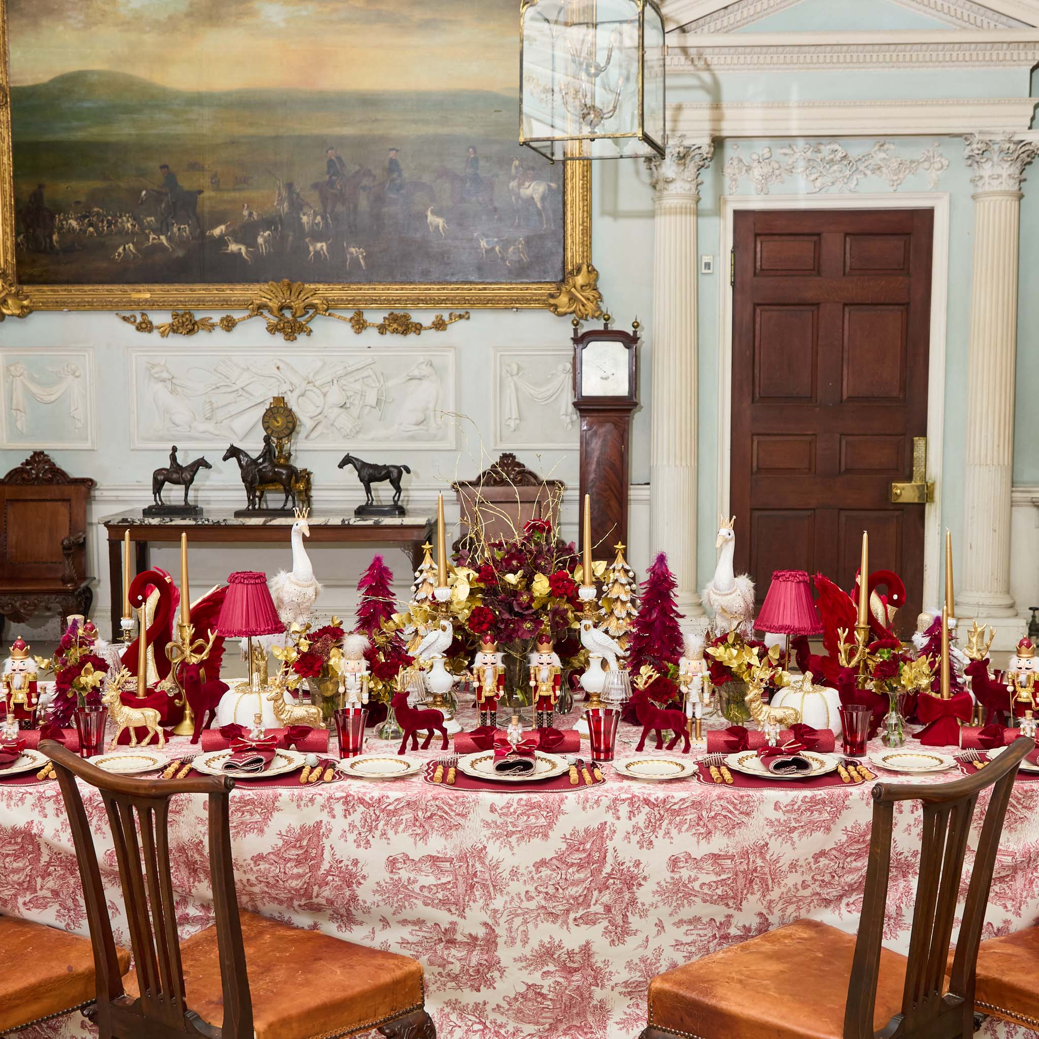 Decorative christmas dining room with a long table set for a formal meal, featuring red and gold tableware and centerpieces.