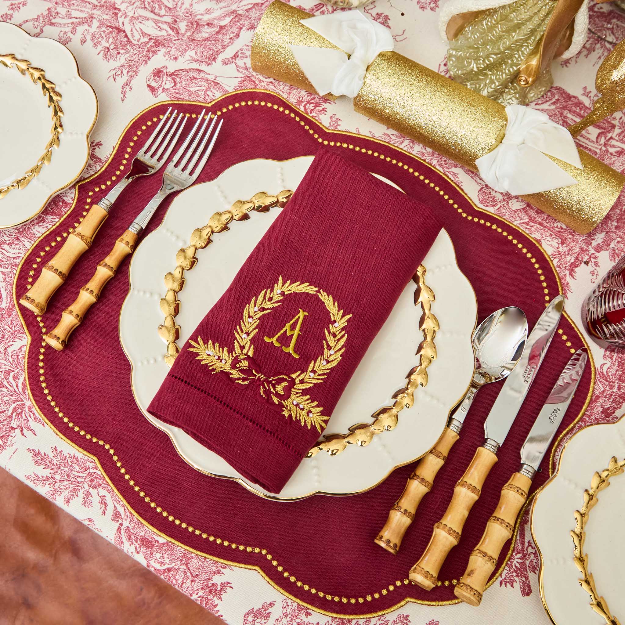 Decorative table setting with red charger, gold cutlery, and a napkin with a monogram.