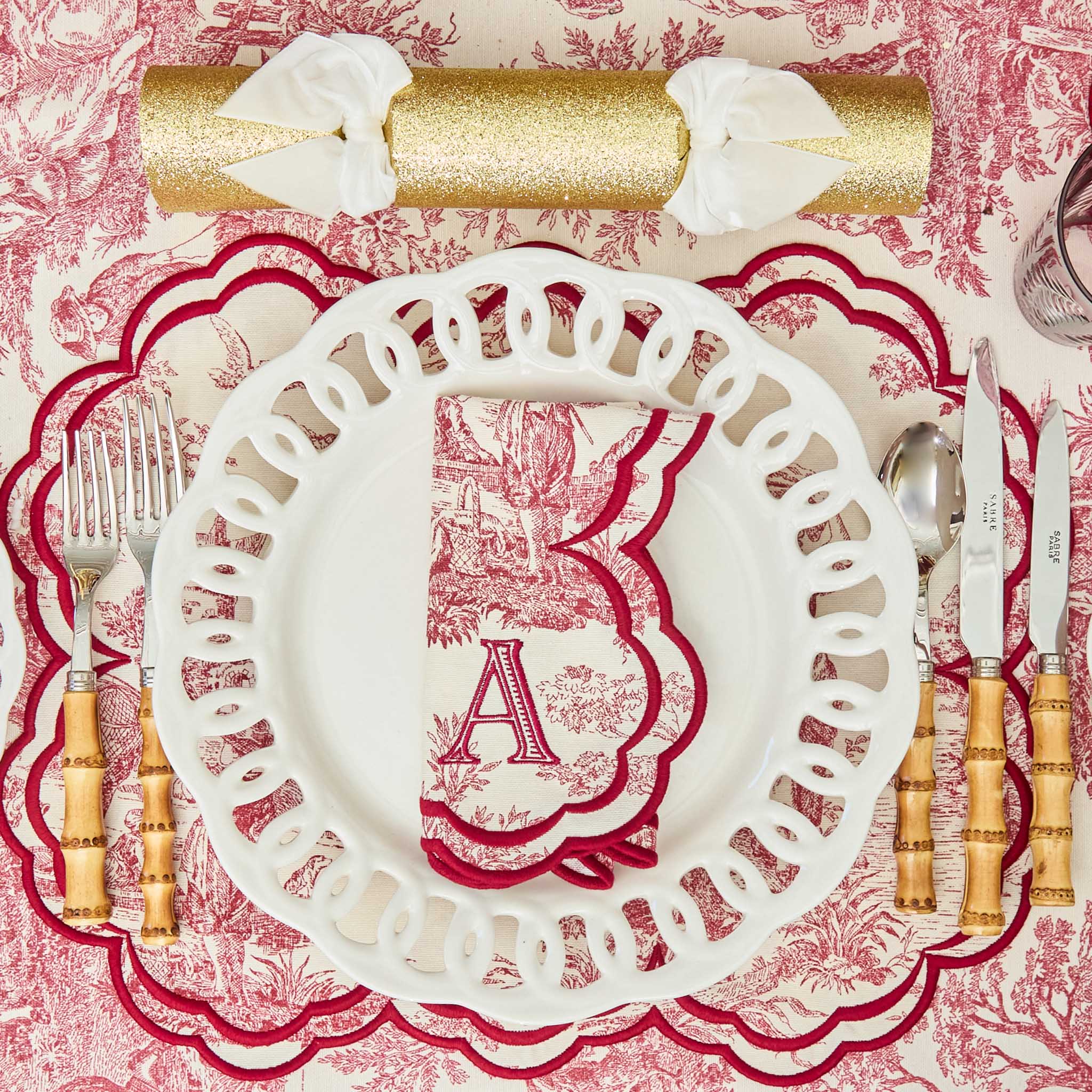 Decorative table setting with a white plate featuring a red letter 'A', gold cutlery, and a red floral patterned tablecloth.