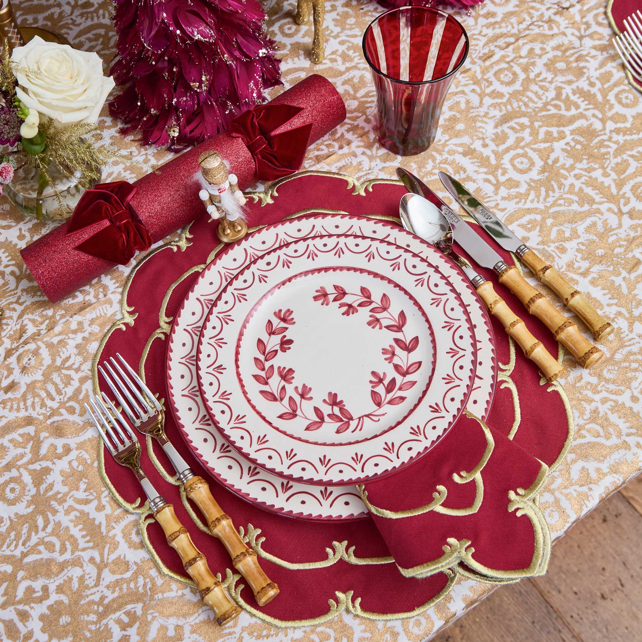 Decorative Christmas table setting with red and gold plates, cutlery, and napkins on a patterned tablecloth.
