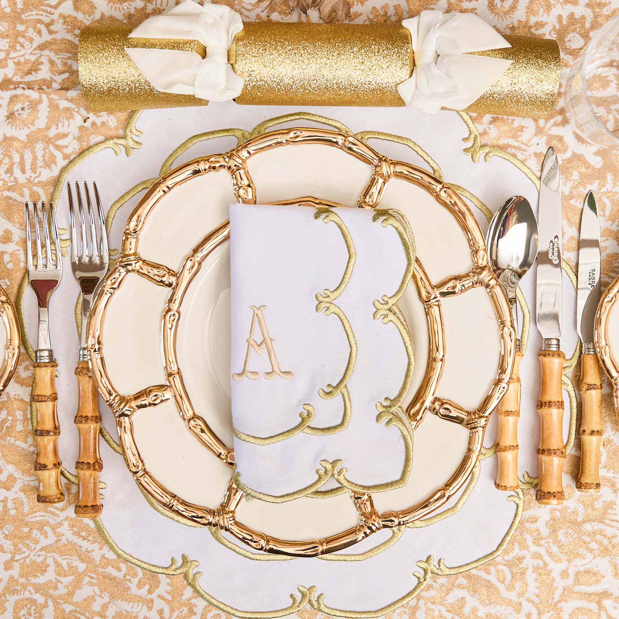 Decorative table setting with gold charger plate, cutlery, and napkin on a patterned tablecloth.
