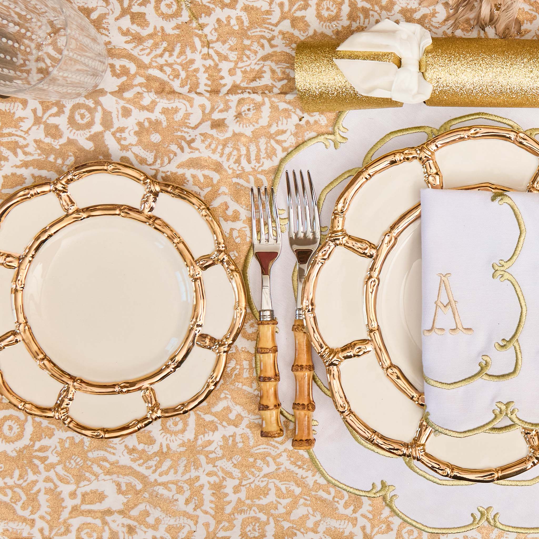Decorative table setting with gold-rimmed plates, cutlery, and a monogrammed napkin on a patterned tablecloth.